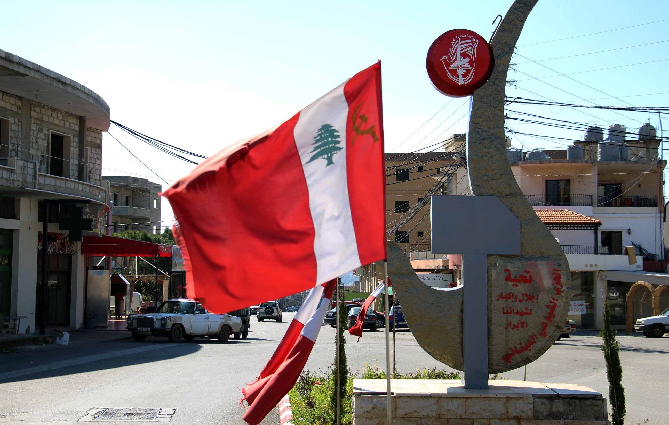 L'image représente un paysage urbain ensoleillé au Liban. On y voit un drapeau libanais flottant au vent, avec une grande emblème en métal en arrière-plan. Cet emblème est orné de symboles et de signification culturelle, illustrant probablement l'identité locale ou nationale. Les bâtiments autour semblent typiques de la région, avec des lignes électriques visibles et un ciel clair en arrière-plan.