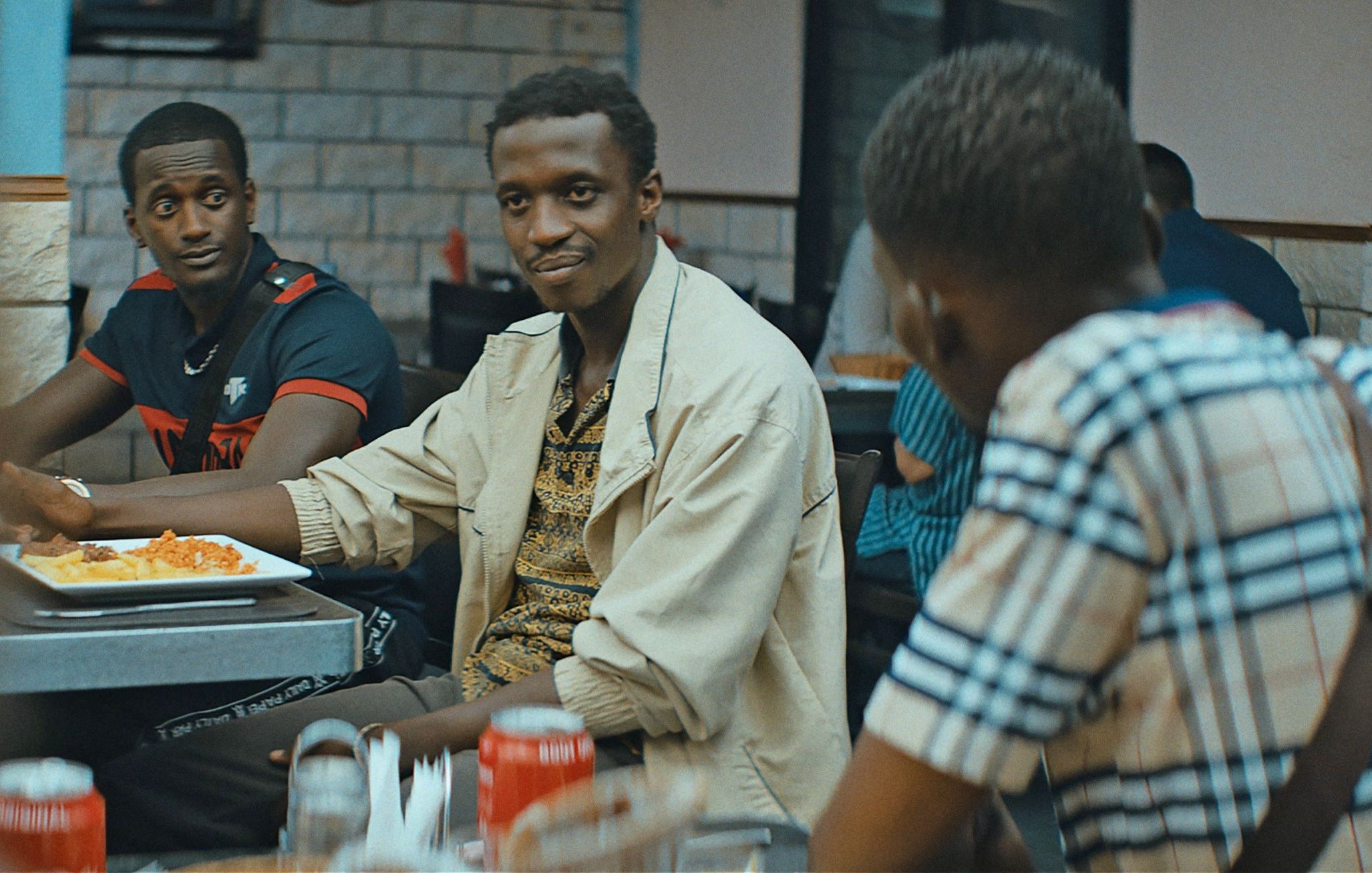 L'image montre un groupe de personnes assises dans un restaurant. Au premier plan, un homme souriant, portant une chemise claire et un motif coloré, semble engager une conversation. Deux autres hommes sont également visibles : l'un avec un maillot noir et rouge, et l'autre portant une chemise à carreaux. Les tables sont garnies de plats de nourriture, et l'ambiance est détendue, typique d'un moment convivial entre amis.