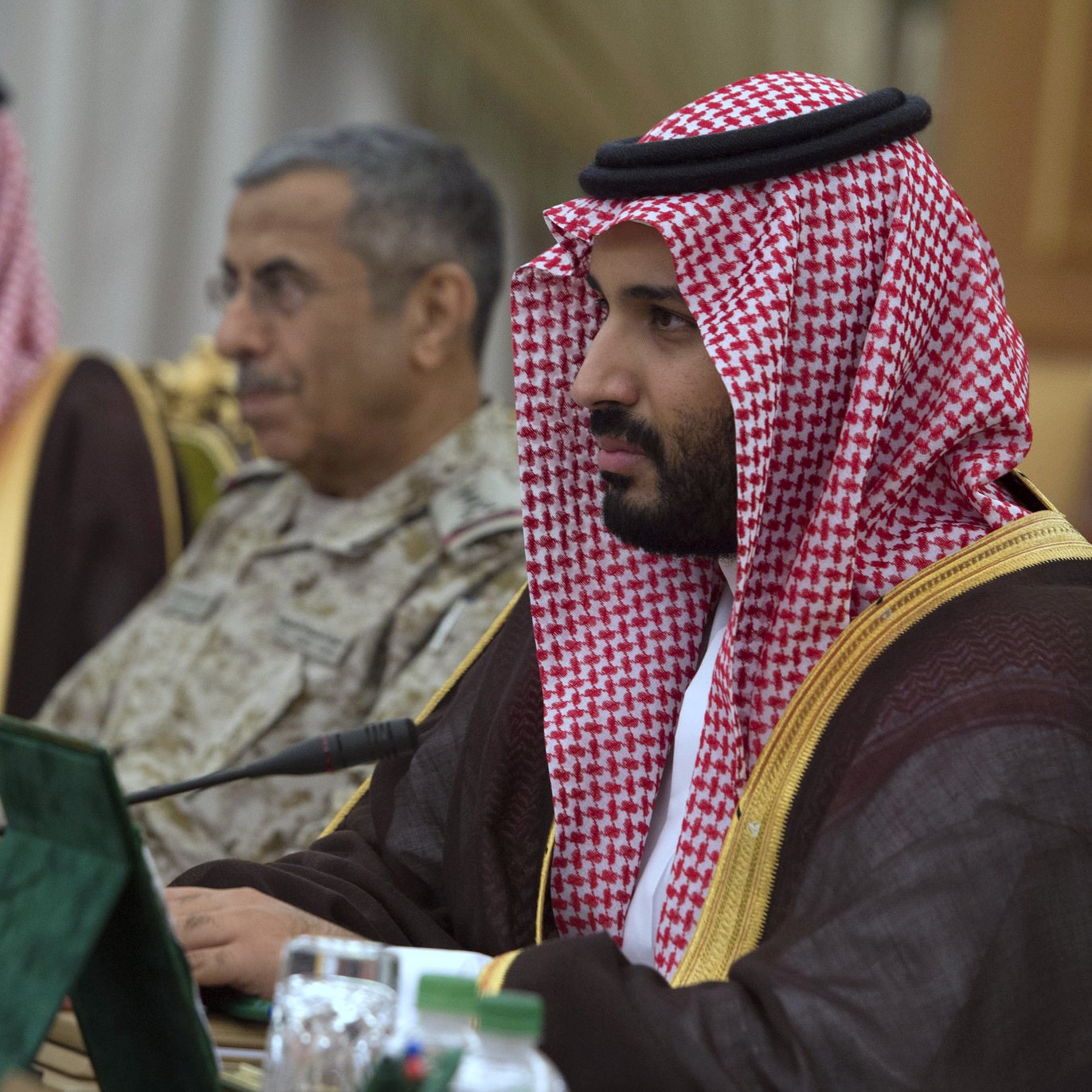 The image shows a group of individuals in formal attire, seated around a table. The man in the foreground is dressed in traditional Saudi attire, wearing a red and white checkered headscarf (ghutrah) and a long brown robe. He appears focused, looking ahead. In the background, another man in military attire is also present, along with other officials. The setting appears to be a formal meeting or conference, indicated by the presence of flags and documents on the table.