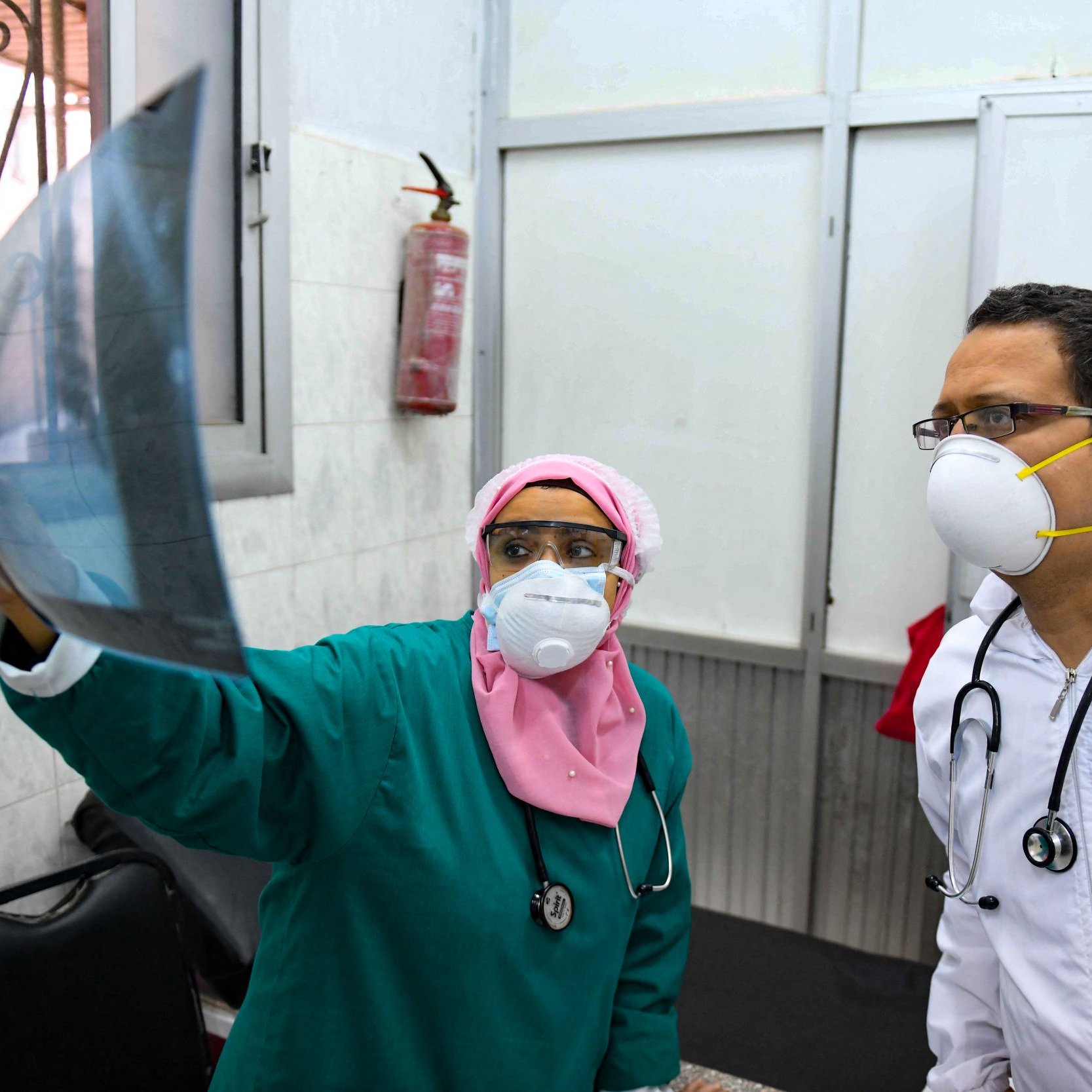 تظهر في الصورة مهنية طبية ترتدي زيًا طبيًا أخضر وغطاء رأس ورديًا، وتفحص صورة أشعة. إلى جانبها، يوجد طبيب يرتدي سترة بيضاء وقناع وجه وقناع تنفس. كلاهما يبدوان مهتمين بالمعلومات الموجودة في صورة الأشعة، ويظهر في الخلفية جدار مكتبي ومعدات طبية.