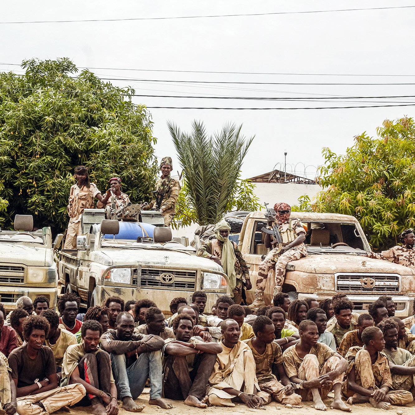 L'image montre un groupe de personnes assises au sol, semblant fatiguées et dans des vêtements usés, tandis qu'elles sont entourées de plusieurs véhicules tout-terrain. À l'arrière-plan, on peut voir des arbres et des plantes, suggérant un environnement rural. Certains individus semblent garder les véhicules, et l'atmosphère générale donne une impression de tension ou de détresse.