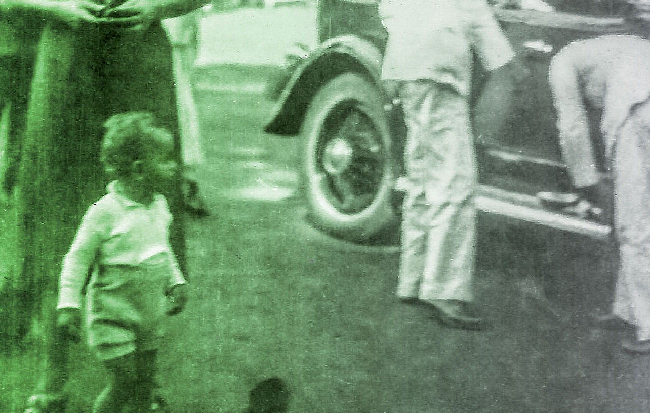 L'image montre un groupe de personnes en extérieur, probablement dans un cadre informel ou festif. On peut voir un enfant en bas à gauche, habillé en short, qui semble jouer. Des adultes, qui peuvent être des membres de la famille ou des amis, se tiennent près d'une voiture vintage, ce qui ajoute une touche rétro à la scène. L'atmosphère semble conviviale et joyeuse. Les vêtements et le style des personnages évoquent une époque passée.