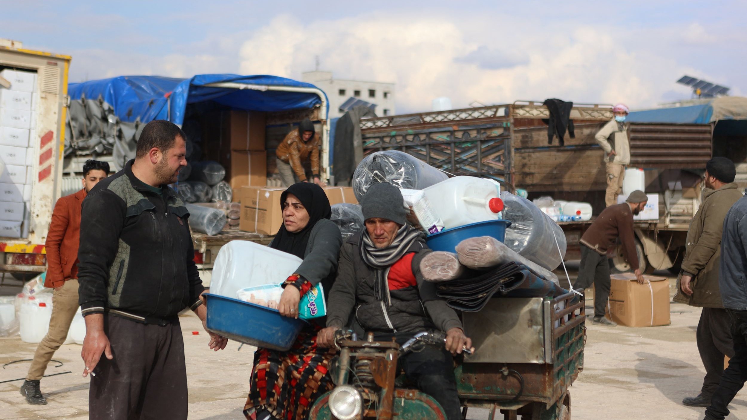 الصورة تظهر تجمعاً في منطقة يبدو أنها مكان لتوزيع المساعدات. يوجد رجل وامرأة يحملان بعض الأغراض، بينما يجلس رجل آخر على عربة صغيرة تحمل زجاجات ومحتويات أخرى. تتواجد خلفهم مجموعة من الأشخاص الذين يعملون على تحميل وتفريغ الأغراض من الشاحنات. السماء تبدو ملبدة بالغيوم، مما يعطي انطباعًا عن الأجواء المحيطة.