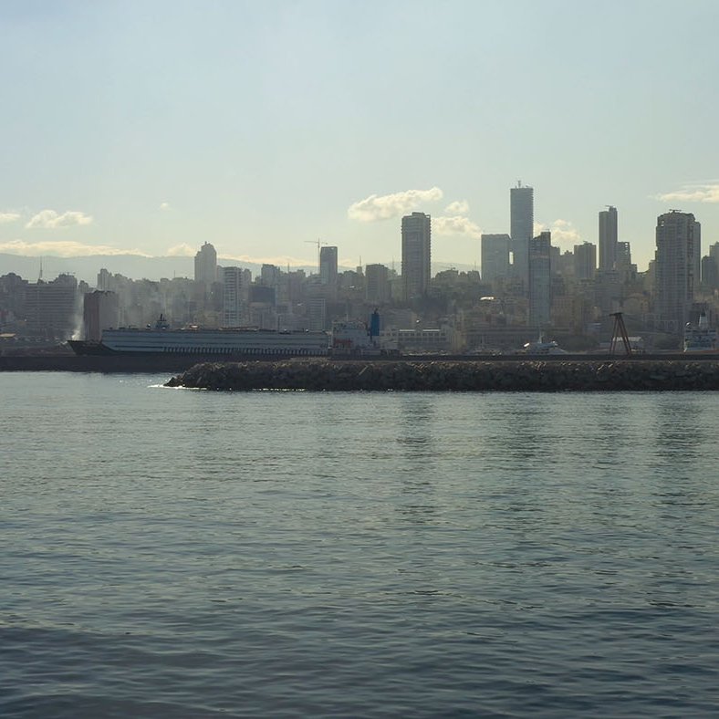 تظهر الصورة منظراً ساحلياً لمدينة حديثة، حيث يمتد الأفق في الخلفية مع مباني شاهقة. السطح المائي هادئ ويعكس ضوء الشمس، مما يخلق جوًا هادئًا. يبدو أن هناك سفينة راسية بالقرب من الميناء، بينما تظهر السحب الخفيفة في السماء فوق المباني. الألوان تبدو دافئة مع ظهور ضباب خفيف يعطي شعورًا بمسافة وتأمل في مشهد المدينة.