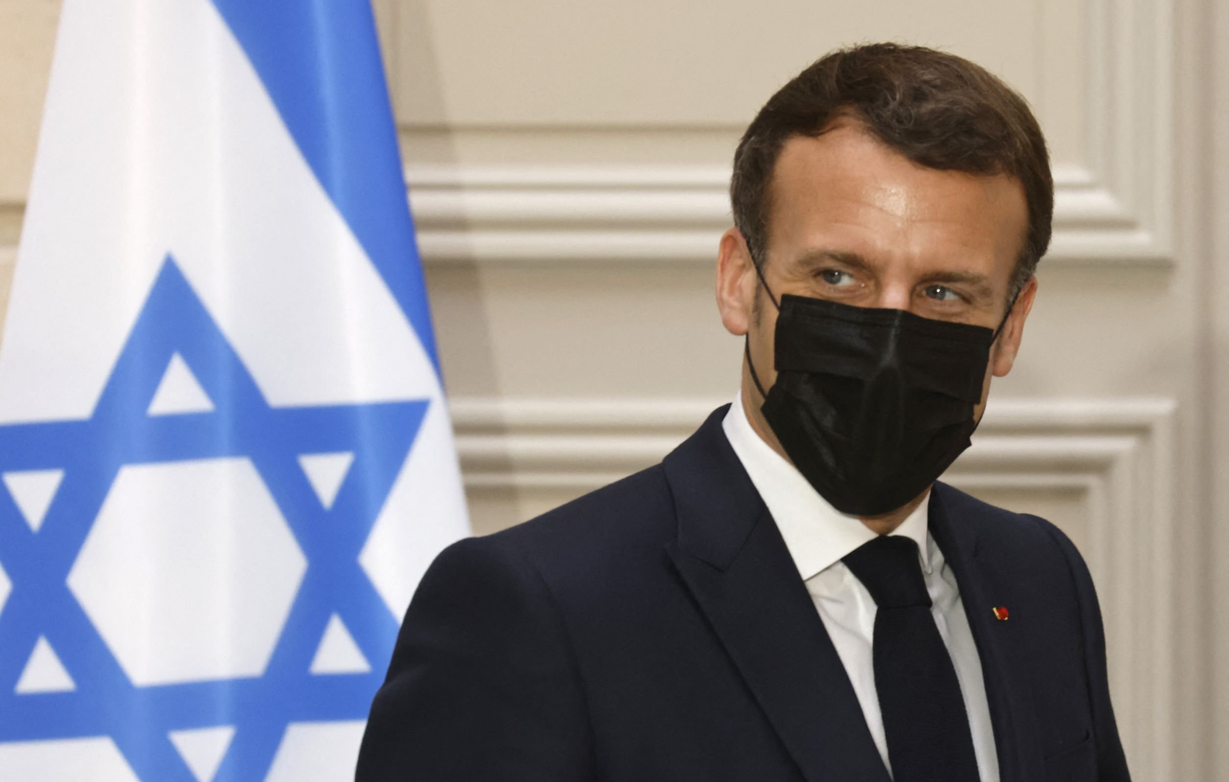 En la imagen se puede ver a un hombre con un traje oscuro y corbata, que lleva una mascarilla negra. Está de perfil y parece estar en un evento oficial. Al fondo hay una bandera con la estrella de David, indicando que el contexto puede estar relacionado con Israel. La expresión del hombre es seria y concentrada.