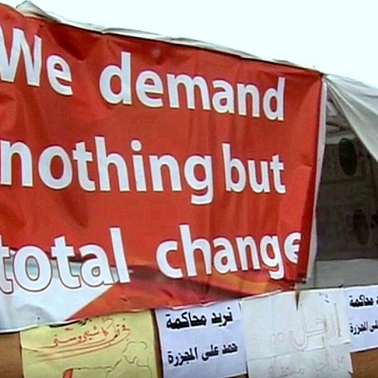 L'image montre une grande pancarte rouge avec le texte "We demand nothing but total change" en anglais, ce qui signifie "Nous exigeons rien d'autre qu'un changement total". En dessous de cette pancarte, on peut voir plusieurs autres affiches, probablement en arabe, qui expriment également des demandes ou des revendications. L'ensemble de la scène évoque un lieu de manifestation ou de protestation, suggérant des aspirations à un changement significatif.