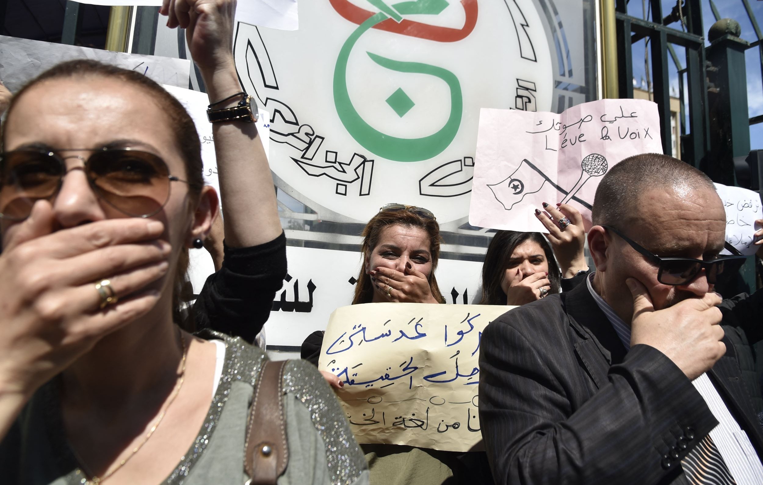 L'image montre une manifestation avec des personnes tenant des pancartes. Elles portent toutes la main sur la bouche, symbole de protestation ou de silence forcé. On peut voir des messages écrits sur les pancartes, qui semblent exprimer des revendications ou des critiques. Les manifestants sont sérieux et déterminés, reflétant un fort engagement pour leur cause. En arrière-plan, il y a un logo ou un symbole en rapport avec le mouvement.