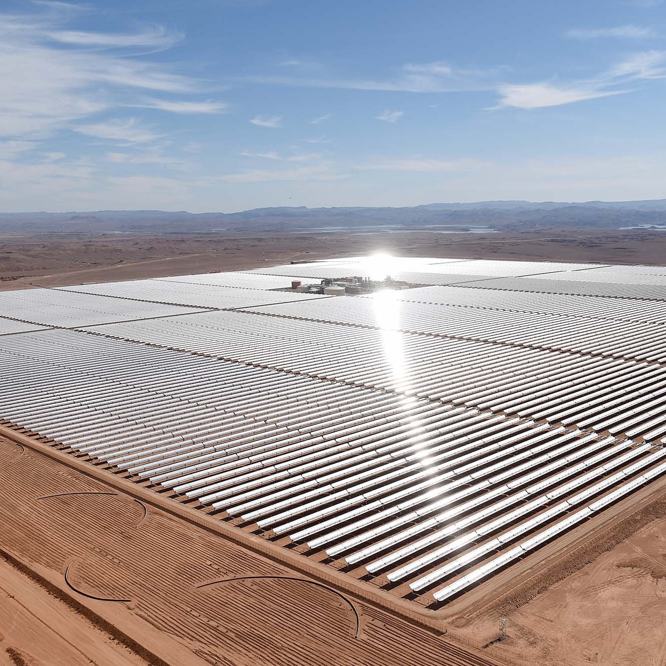 L'image montre une vaste installation de panneaux solaires, s'étendant sur une grande surface de terrain désertique. Les panneaux sont disposés en rangées régulières, reflétant la lumière du soleil. On peut apercevoir des collines à l'arrière-plan, et le ciel est clair avec quelques nuages. L'ensemble donne une impression de modernité et de durabilité, soulignant l'importance des énergies renouvelables dans le paysage contemporain.