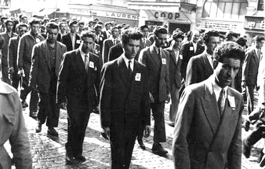 L'image montre un groupe d'hommes marchant ensemble dans une rue pavée. Tous portent des costumes et semblent sérieux. Ils avancent en rang, avec des badges sur leur veste. En arrière-plan, on peut distinguer quelques bâtiments et enseignes, ce qui donne un sens d'époque à la scène. L'atmosphère peut paraître solennelle, suggérant possiblement une manifestation ou un événement organisé.