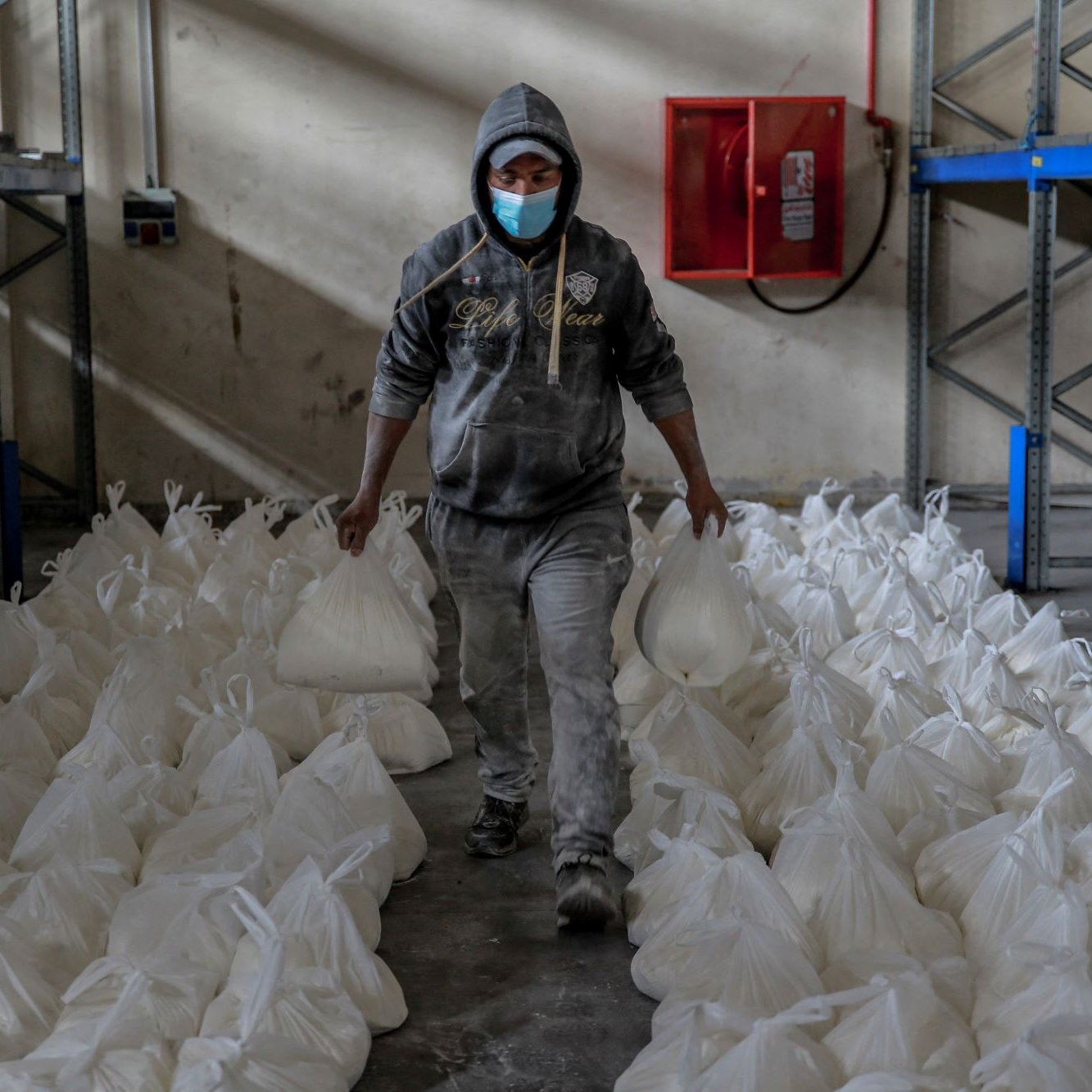 در این تصویر، یک مرد با پوشش خاکستری و کلاهی بر سر، در حال حرکت در یک محل انبار است. او یک ماسک به صورت دارد و در دو دست خود کیسه‌های سفیدی را حمل می‌کند. اطراف او ردیف‌هایی از کیسه‌های مشابه به طور منظم روی زمین قرار دارند. دیوارها و قفسه‌های فلزی در پس‌زمینه قابل مشاهده هستند که به نظر می‌رسد برای نگهداری کالاها استفاده می‌شوند. نور کمی در فضا وجود دارد و جو عمومی، فعالیتی را در زمینه توزیع یا انبارداری نشان می‌دهد.