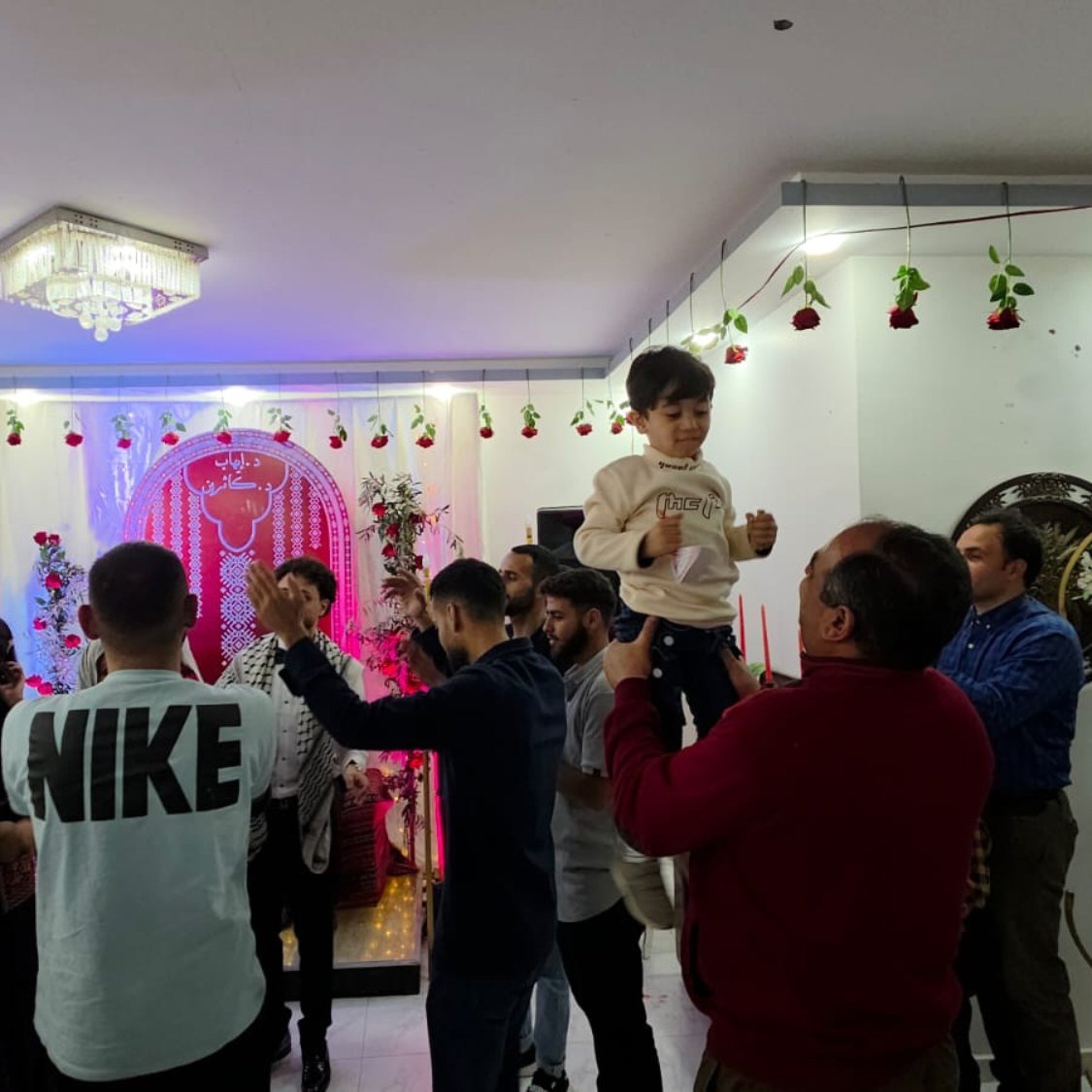 فضای شاد، مردم در حال رقص، تزئینات گل و نورپردازی رنگارنگ، یک کودک در آغوش بزرگترها.