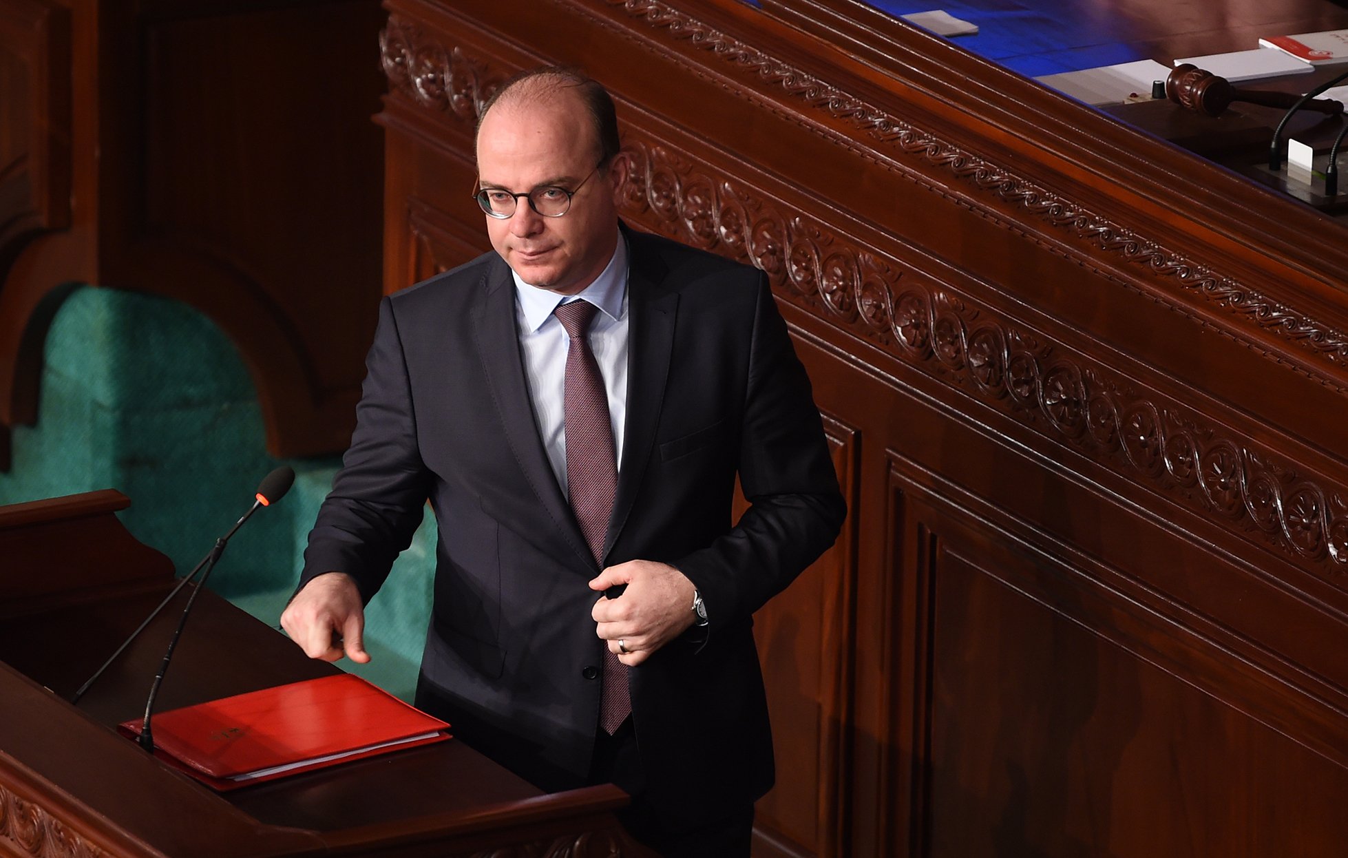 L'image montre un homme en costume sombre, portant des lunettes, se tenant près d'un pupitre dans une assemblée ou un parlement. Il semble être en train de s'adresser à l'assemblée ou de faire une déclaration informative, avec un dossier rouge posé sur le pupitre. Le décor est constitué de boiseries et de sièges verts, typiques d'un environnement institutionnel.