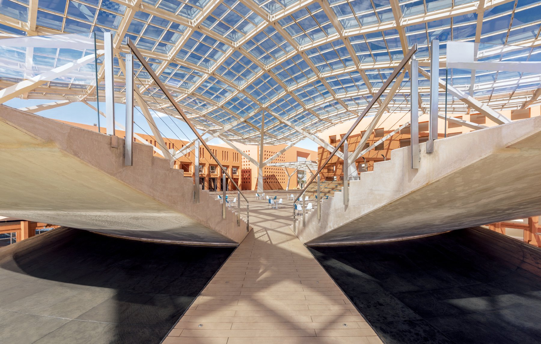 L'immagine mostra un'architettura moderna caratterizzata da una struttura ampia e luminosa. La copertura in vetro permette alla luce naturale di filtrare, creando un ambiente aperto e arioso. Le pareti laterali presentano un design minimalista, con elementi di legno e cemento che si combinano armoniosamente. Al centro, una passerella in legno collega le diverse aree, accentuando una sensazione di continuità e apertura nello spazio. La scena è caratterizzata da linee pulite e forme geometriche, suggerendo un design innovativo e funzionale.