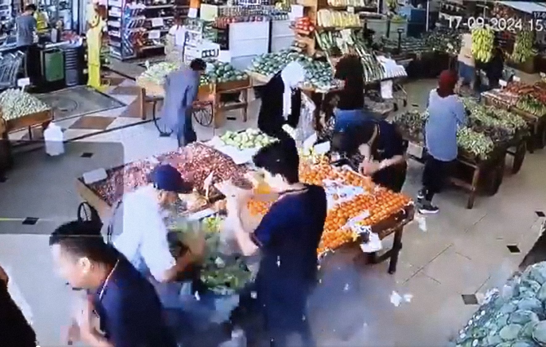 L'immagine mostra l'interno di un negozio di alimentari, probabilmente una fruttivendolo. Ci sono diverse persone che si muovono tra le corsie, mentre vari tipi di frutta e verdura sono esposti su tavoli e scaffali. Le persone sembrano occupate a scegliere e acquistare prodotti freschi. La scena è vivace, con molti colori provenienti dai frutti esposti, come agrumi e verdure di vario tipo. La disposizione degli oggetti suggerisce un'ambientazione ordinata e ben fornita.