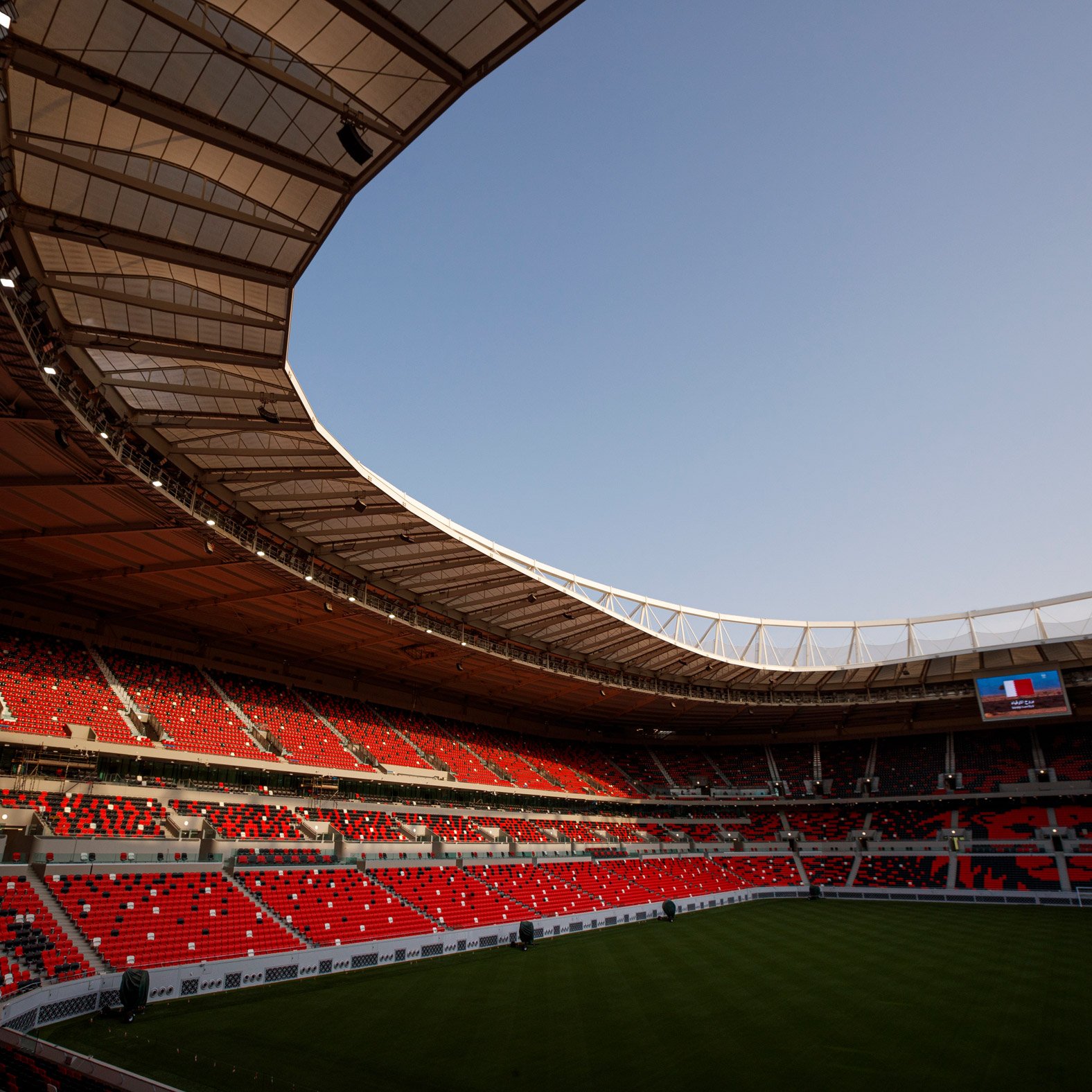 L'image montre un stade moderne avec des sièges rouges et une large toiture. La pelouse est bien entretenue, et on peut apercevoir un écran géant à une extrémité. Le ciel est dégagé, indiquant une belle journée. L'intérieur du stade semble prêt à accueillir un événement, avec une atmosphère calme et anticipative.