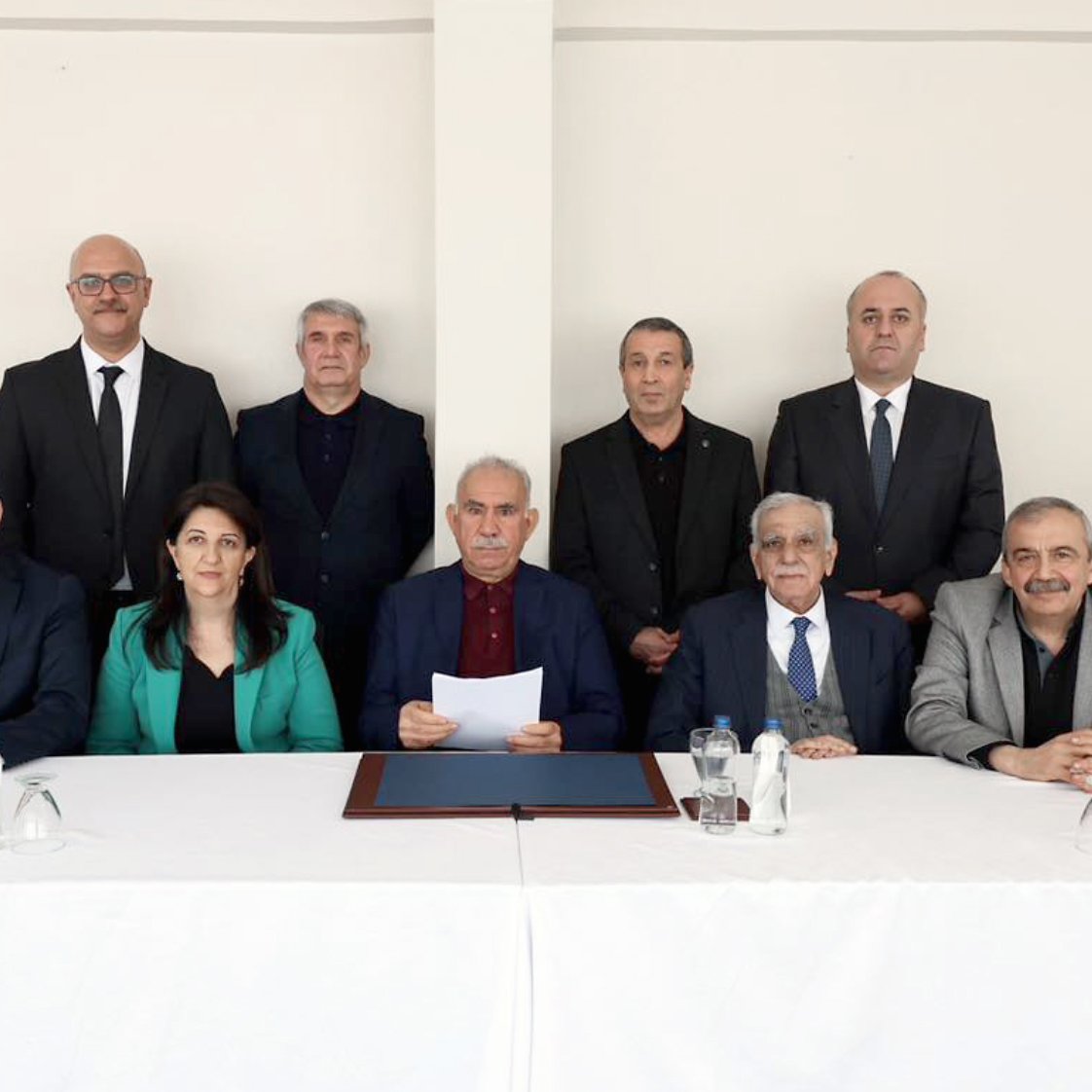 L'image montre un groupe de douze personnes assises derrière une table blanche. Il y a des hommes et des femmes, tous vêtus de costumes et de tenues professionnelles. L'ambiance semble formelle, et une personne au centre tient un document.