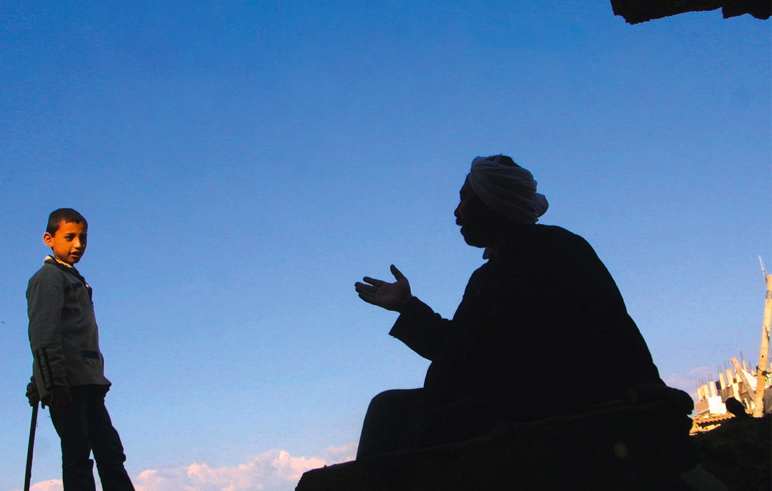 L'image montre deux silhouettes. À gauche, un garçon se tient debout, regardant vers l'autre personne. À droite, un homme est assis, en train de parler, ses bras gesticulant. Le fond est un ciel bleu, ce qui crée un fort contraste avec les silhouettes noires. L'environnement semble brut, avec des débris visibles, indiquant un cadre peut-être urbain ou une zone touchée par des événements difficiles.
