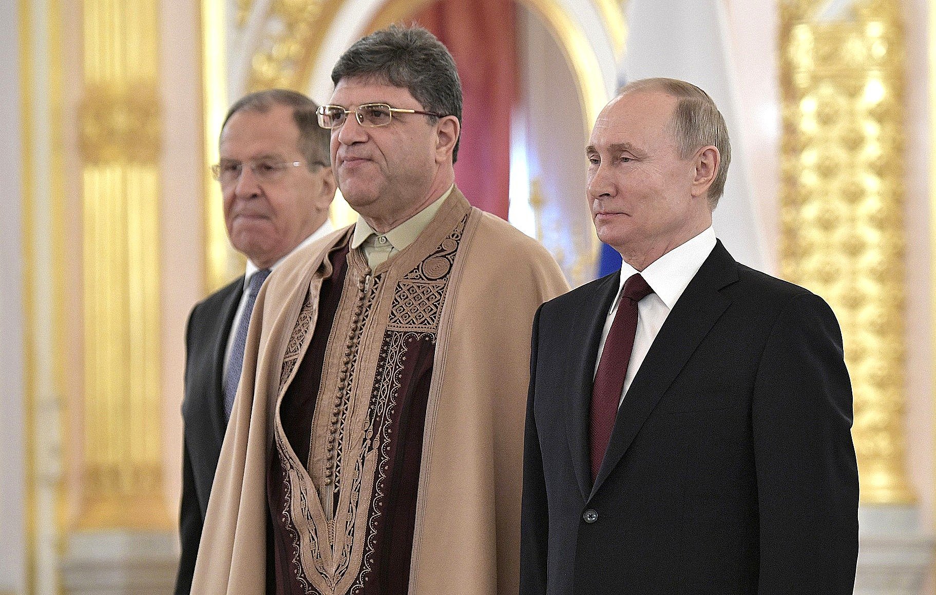 The image shows three men standing together in what appears to be an official setting, likely a ceremonial event. The man on the left is dressed in a suit, while the man in the middle is wearing traditional attire, featuring intricate patterns. The man on the right, who is likely a well-known political figure, is also dressed in a formal suit. The background is ornate, suggesting a formal and significant atmosphere.