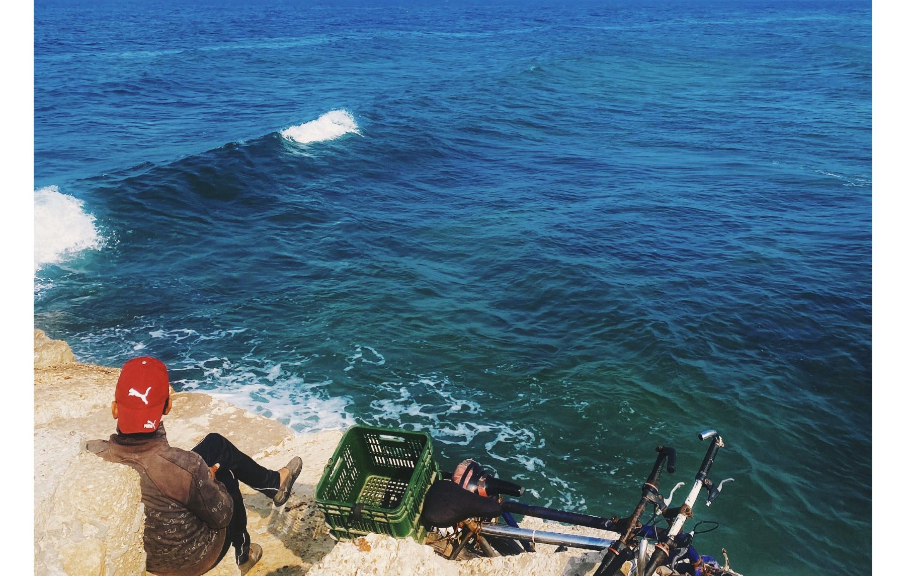 Un homme assis sur des rochers, face à une mer calme et bleue sous un ciel dégagé.