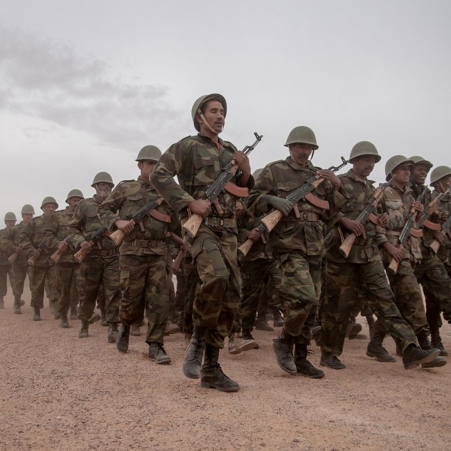 L'image représente un groupe de soldats marchant en formation sur un chemin poussiéreux. Ils sont habillés en tenue militaire camouflée et portent des casques. Chaque soldat tient un fusil à la main. L'atmosphère semble sérieuse, et le ciel est nuageux, suggérant peut-être des conditions climatiques difficiles. En arrière-plan, on aperçoit une ligne de végétation ou d'arbres, ajoutant à l'impression d'un environnement militaire.