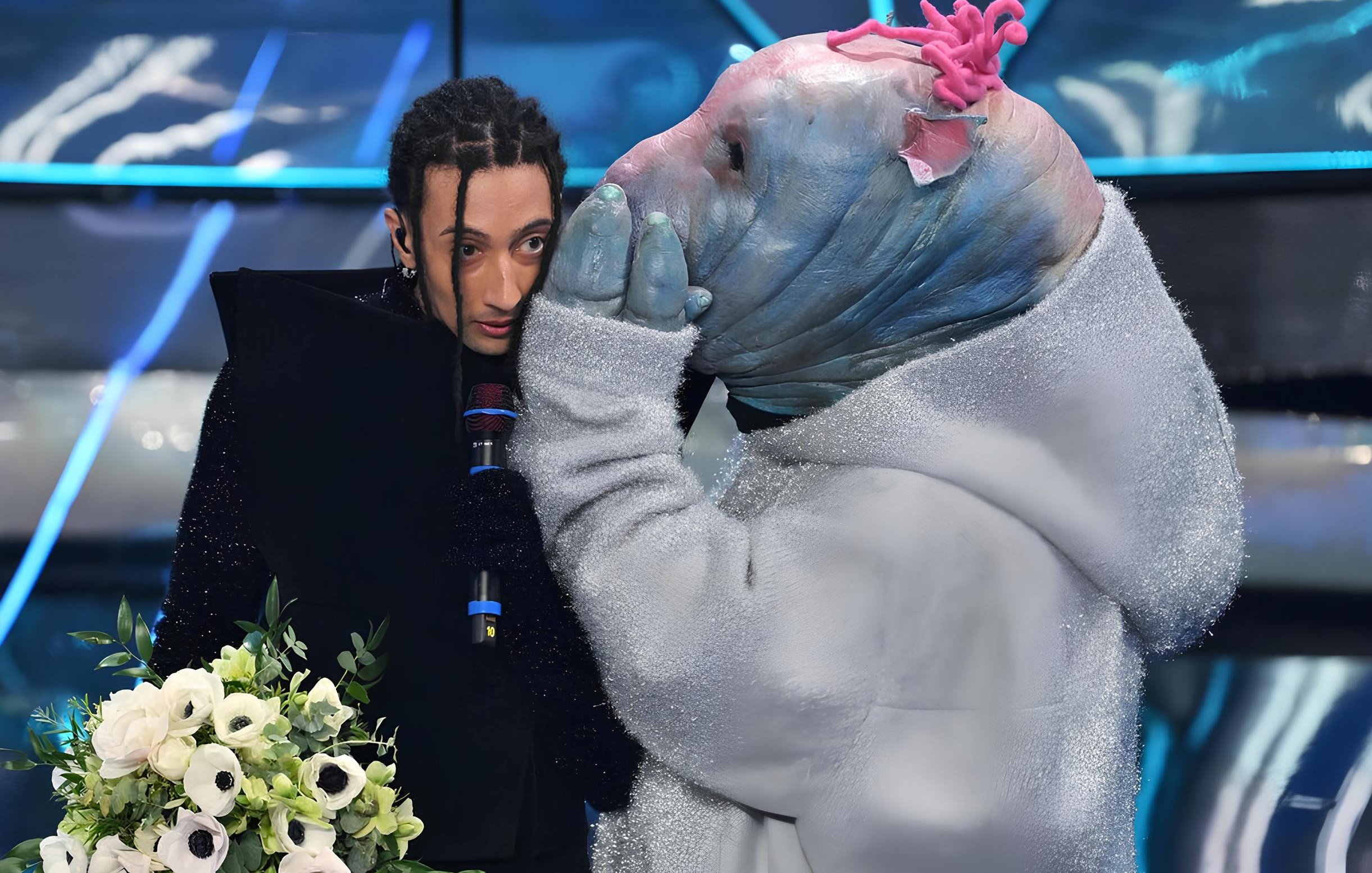 L'image montre une scène d'un spectacle où une personne au style vestimentaire unique, tenant un bouquet de fleurs, semble être en conversation avec une autre personne déguisée dans un costume original. Le costume ressemble à un animal aquatique, avec une tête d'apparence bleue et des détails colorés. L'ambiance semble dramatique ou émotive, avec un éclairage qui accentue les éléments de la scène.