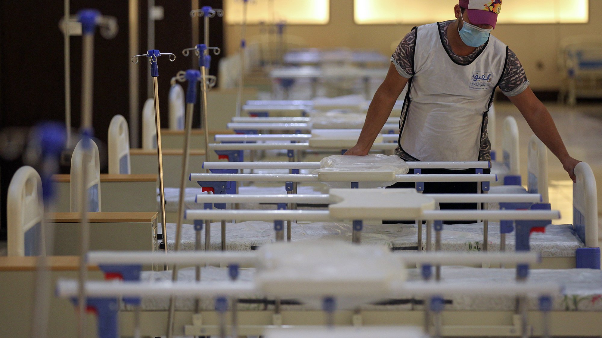 ترتدي الصورة مشهداً داخل مرفق صحي، حيث يظهر شخص واحد يرتدي كمامة وقميصاً. الشخص يركز على سرير طبي بين عدة أسرّة مرتبة بشكل منظم في غرفة واسعة مضاءة بشكل جيد. تظهر في الخلفية أعمدة ومعدات طبية، مما يوحي بأن المكان يُستخدم لعلاج المرضى. يتميز الجو بالهدوء والترتيب.