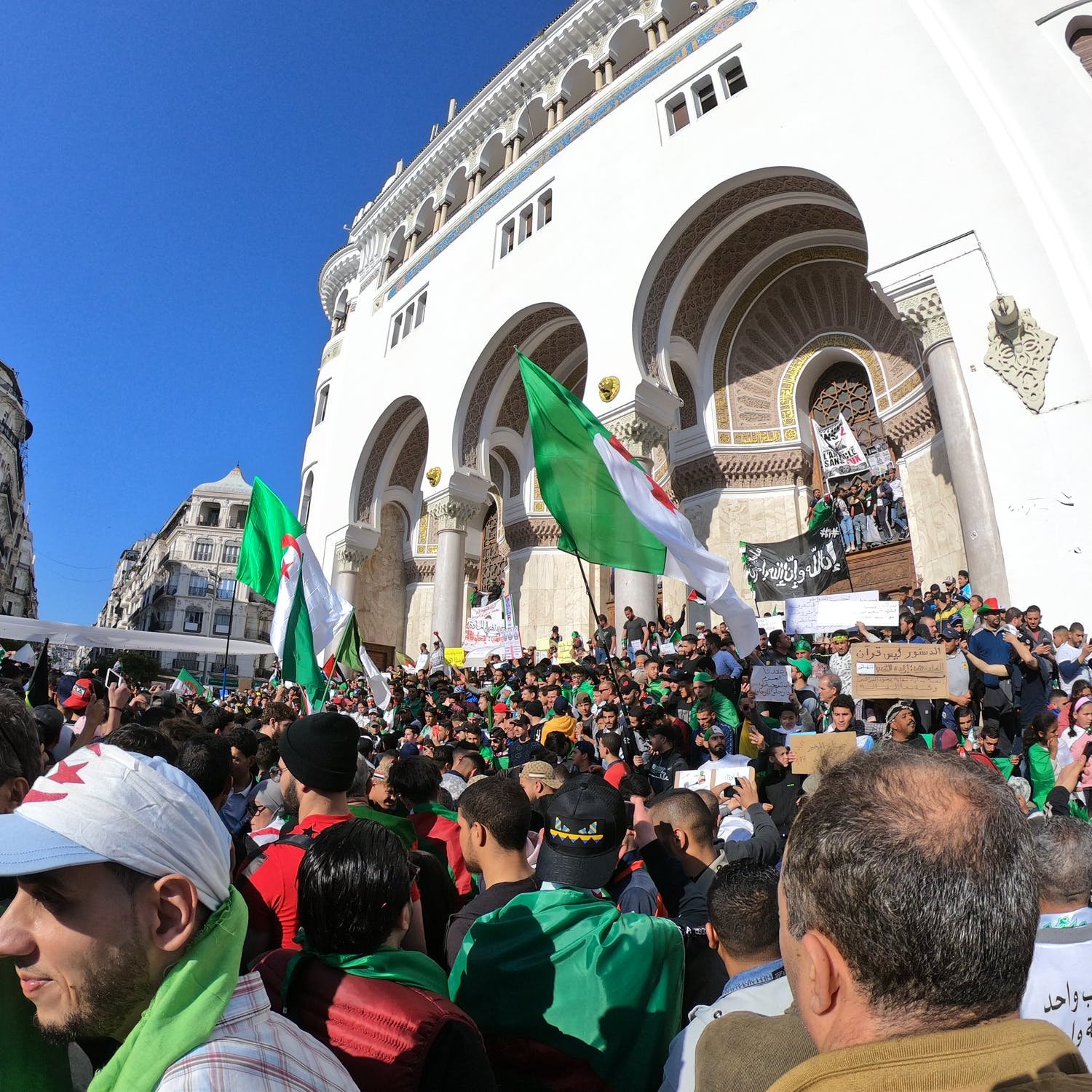 L'image montre une grande foule rassemblée devant un bâtiment avec une architecture traditionnelle. Les manifestants brandissent des drapeaux algériens et des pancartes, exprimant probablement des revendications politiques ou sociales. La scène est en plein air, sous un ciel bleu, et il y a une atmosphère de mobilisation collective, avec des personnes de tous âges vertes et rouges.