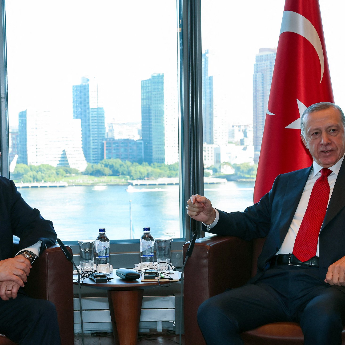 L'image montre deux hommes en discussion dans un cadre professionnel. À gauche, il y a un homme avec une cravate rouge, assis sur une chaise, regardant l'autre homme. À droite, l'autre homme, également en cravate, semble parler et gesticuler. En arrière-plan, on aperçoit des gratte-ciel et une vue sur l'eau, suggérant un environnement urbain. Des drapeaux d'Israël et de Turquie sont visibles, indiquant le contexte de cette rencontre.