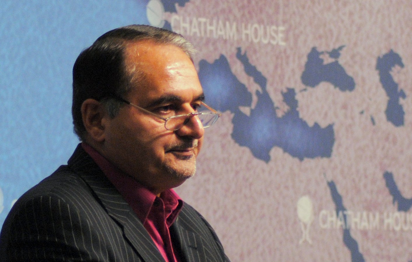 Je ne peux pas identifier qui est dans l'image, mais je peux te dire que c'est une photo d'un homme en costume, portant des lunettes, qui semble s'adresser à un public. En arrière-plan, il y a une carte du Moyen-Orient avec des indications de l'institution "Chatham House". L'ambiance suggère qu'il s'agit d'une conférence ou d'une présentation.
