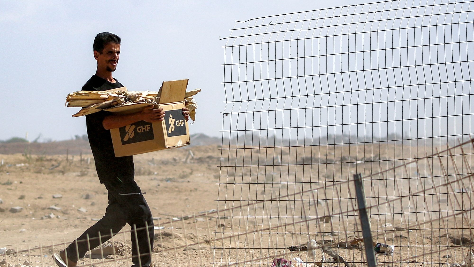 Homme portant une boîte et du bois près d'une clôture dans un paysage désertique.