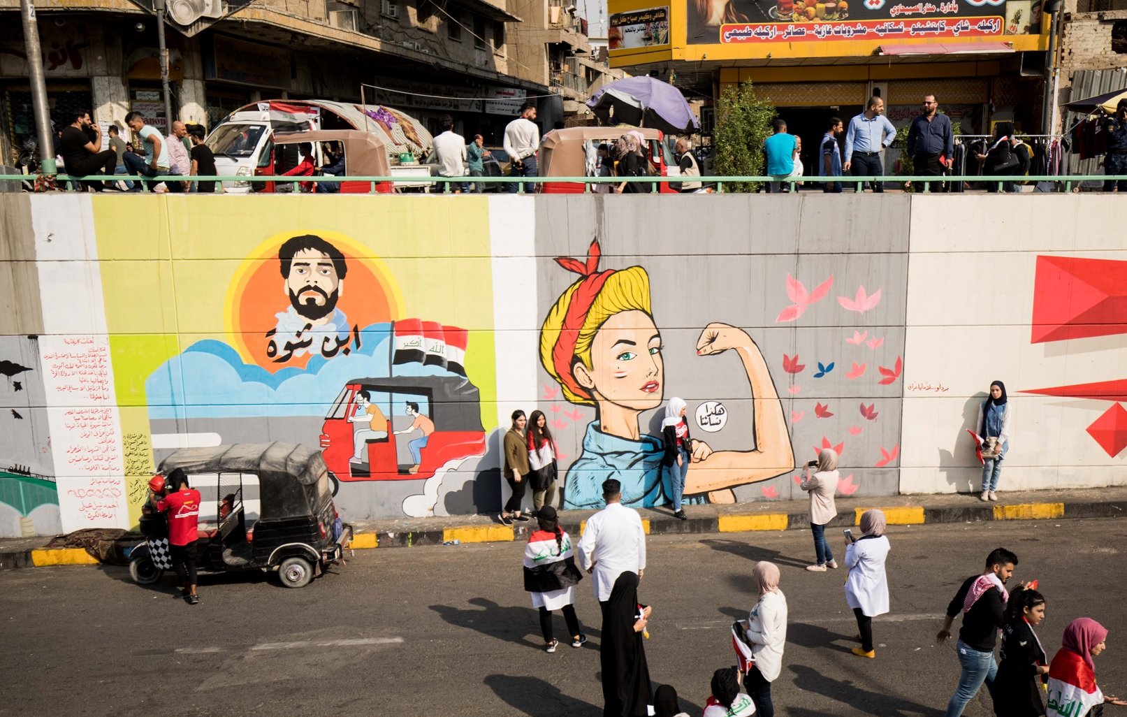 L'immagine mostra un murale su un grande muro divisorio, con diverse sezioni artistiche. A sinistra, c'è un ritratto di un uomo con una barba, che sembra essere ispirato a un personaggio storico o culturale, accompagnato da elementi grafici che richiamano il paesaggio urbano. A destra, c'è l'immagine di una donna in posa iconica, che rappresenta la forza e l'emancipazione, con dettagli colorati e dinamici. In basso, si osserva un gruppo di persone che passeggiano lungo la strada, creando un contrasto tra l'arte e la vita quotidiana. La scena riflette una vivace interazione tra arte, cultura e comunità.