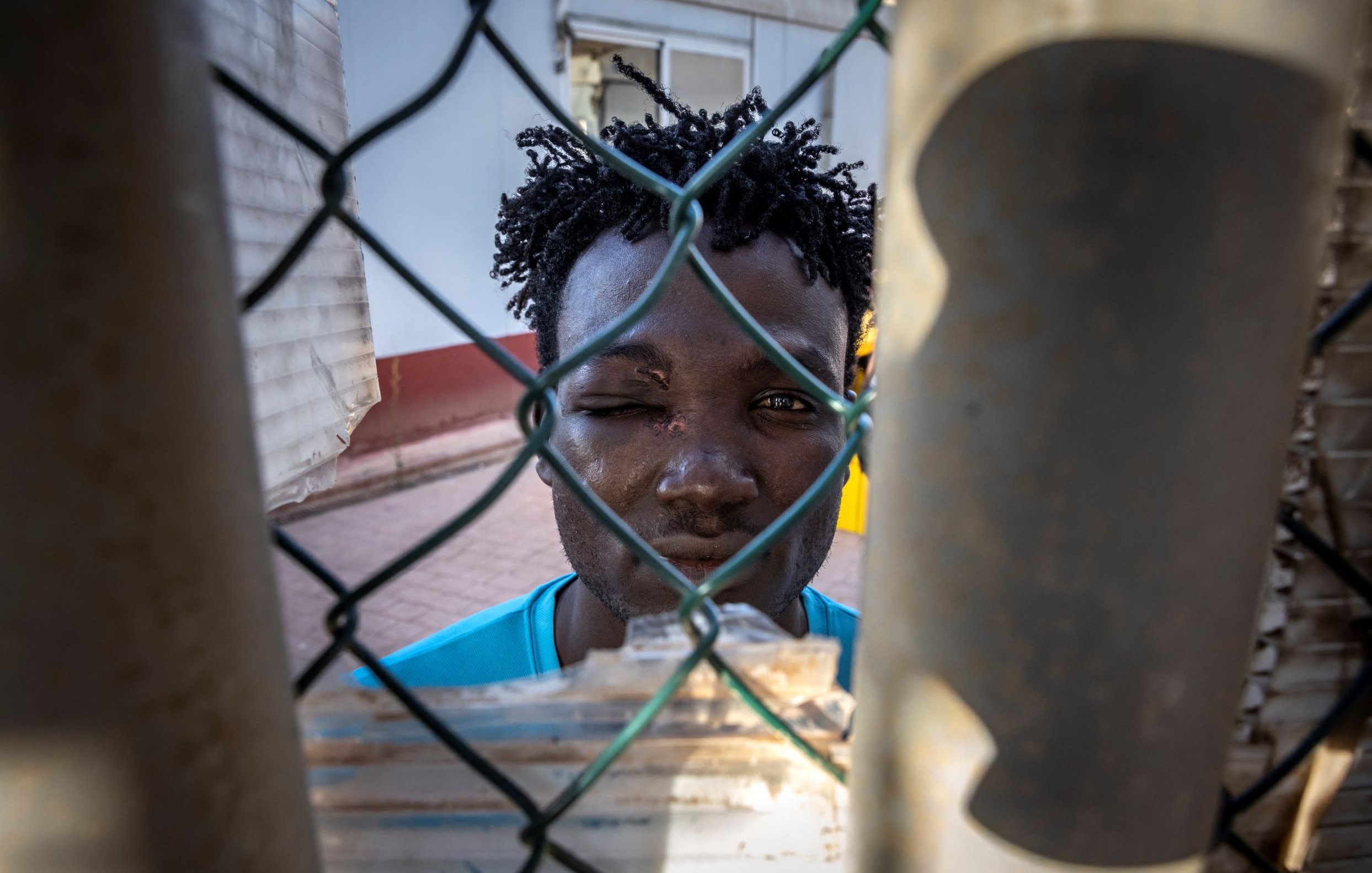 L'immagine mostra un giovane uomo che si affaccia attraverso una rete di recinzione. Ha i capelli ricci e un'espressione seria. Il suo viso è parzialmente coperto da una rete metallica, il che crea un effetto di separazione. Sullo sfondo, si intravede un'architettura semplice. La luce colpisce il suo volto, mettendo in evidenza i dettagli della sua espressione.