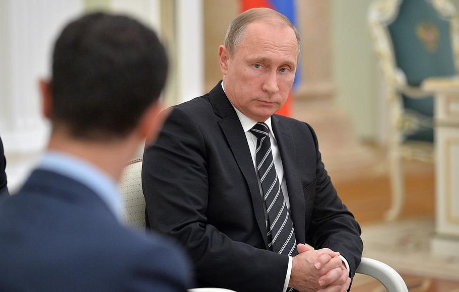 L'image montre un homme assis en costume noir avec une cravate, ayant une expression sérieuse sur le visage. Il semble être en train d'écouter ou de réfléchir pendant une discussion. On peut apercevoir une autre personne au premier plan, de dos, qui ne peut pas être identifiée. L'environnement semble être un bureau officiel, avec des éléments décoratifs typiques d'un cadre institutionnel.