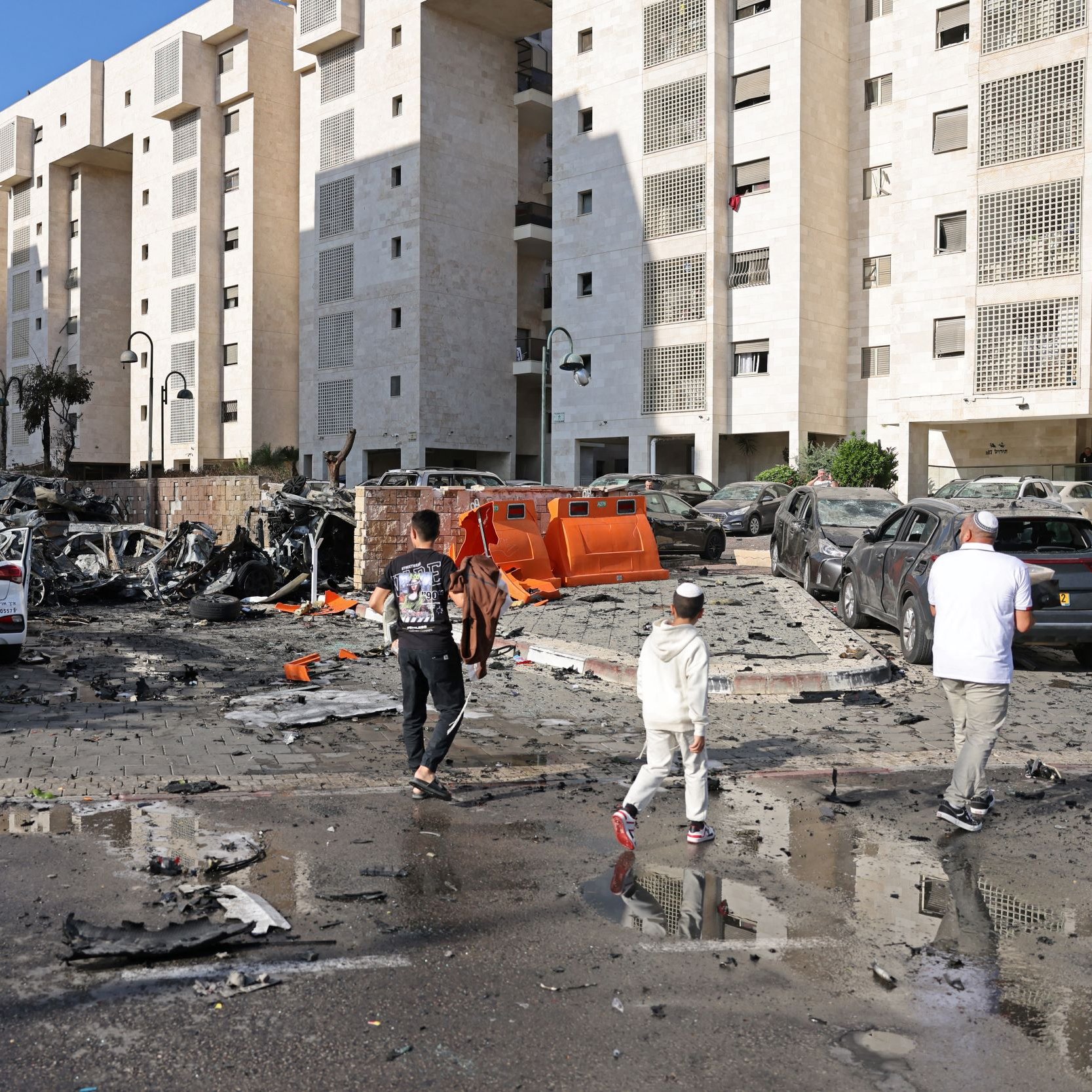 در این تصویر، یک منطقه شهری به نمایش گذاشته شده که به نظر می‌رسد به خاطر یک حادثه انفجاری آسیب دیده است. ساختمان‌ها در دو طرف خیابان قرار دارند و در میان آنها، خودروهای beschädigt و ویران‌شده به چشم می‌خورند. زباله‌ها و قطعات شکسته در سطح زمین پخش شده‌اند و نشانه‌هایی از ویرانی دیده می‌شود. چند نفر در حال قدم زدن در این مکان آسیب‌دیده هستند و به نظر می‌رسد در حال ارزیابی وضعیت هستند. همچنین، آب‌ای که ممکن است بر اثر باران یا دیگر عوامل جمع شده باشد، در بعضی نقاط خیابان دیده می‌شود. آسمان آبی و روشن به نظر می‌رسد.