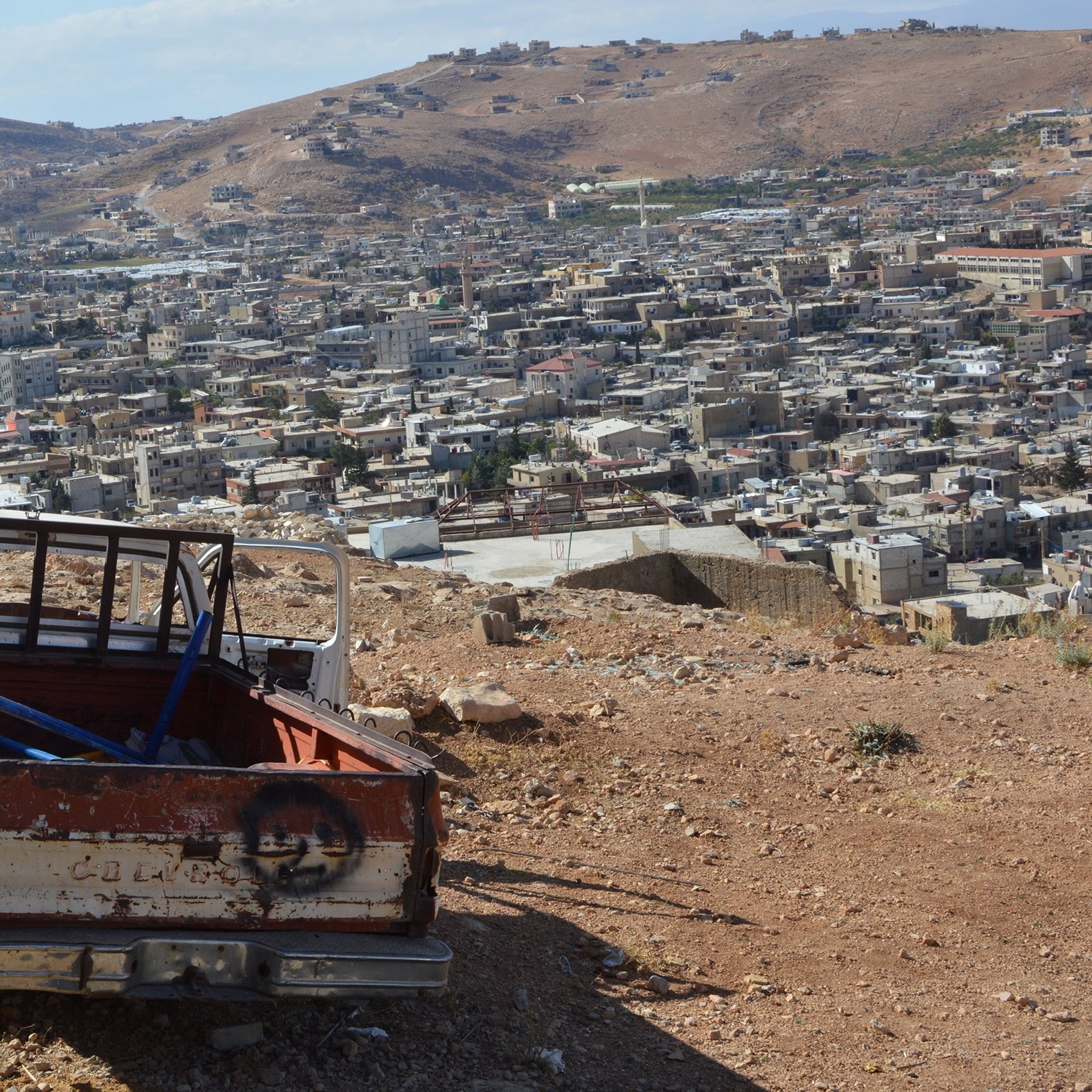 L'image montre une scène de paysage où un ancien véhicule, probablement une camionnette, est abandonné sur une colline. La camionnette est en mauvais état, avec une partie de la carrosserie rouillée et peinte en blanc et rouge. En arrière-plan, on peut voir un village ou une ville avec de nombreuses maisons, s'étendant sur les collines. Le ciel est partiellement nuageux, et le paysage environnant est aride, typique des terrains montagneux.