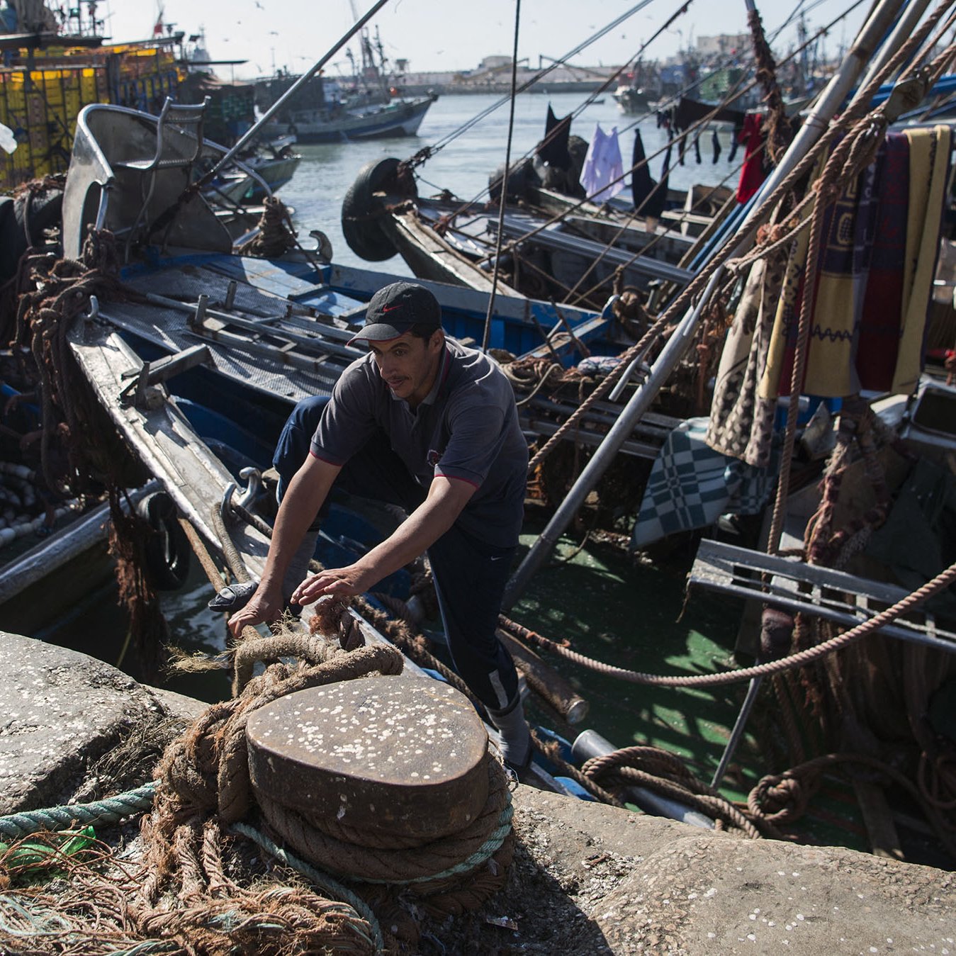 L'image montre un port de pêche animé, avec des bateaux de pêche amarrés. Un homme, vêtu d'un t-shirt et d'une casquette, est visible en train de travailler sur le quai, s'occupant de cordages. En arrière-plan, on peut voir d'autres bateaux et une atmosphère maritime. Le paysage évoque une activité portuaire typique, avec plusieurs éléments liés à la pêche.