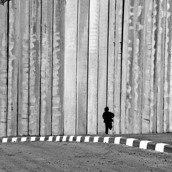 L'image montre un enfant courant le long d'un mur de béton. Le mur est haut et semble imposant, créant une ambiance de séparation ou d'isolement. Le contraste en noir et blanc accentue les textures du mur et la silhouette de l'enfant, qui semble minuscule par rapport à la structure massive. Le sol est désertique avec une route qui tourne légèrement, ajoutant à l'impression de solitude et de mouvement.