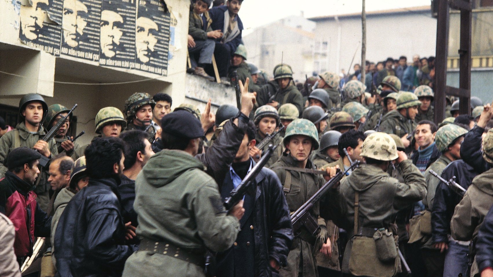 L'image montre un groupe de soldats en uniforme, portant des casques et armés, réunis autour d'une foule. On peut voir des hommes en civil, certains semblent s'opposer aux militaires. En arrière-plan, des affiches sont visibles sur le mur, peut-être représentant des figures politiques ou des messages de protestation. L'atmosphère est tendue et indique un moment de conflit ou de manifestation. Les visages des individus expriment des émotions variées, allant de la détermination à l'inquiétude.