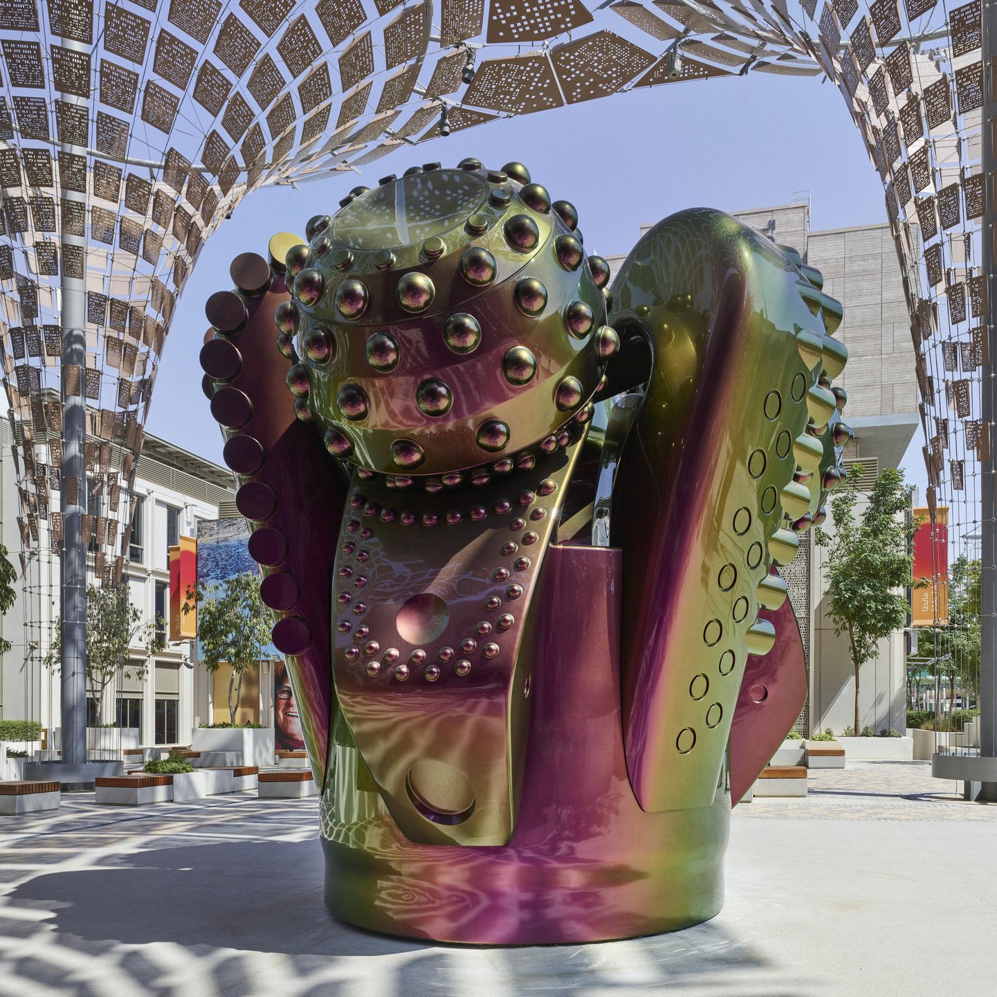 L'image présente une sculpture moderne au design futuriste, composée de formes organiques et géométriques. Les surfaces de la sculpture sont brillantes et colorées, avec des reflets irisés qui passent du vert au violet. Elle semble être placée sous une structure architecturale légère, qui offre de l'ombre et favorise une ambiance artistique. L'environnement autour de la sculpture est minimaliste, avec des arbres et quelques assises, ce qui met en valeur l'œuvre et crée une atmosphère apaisante.