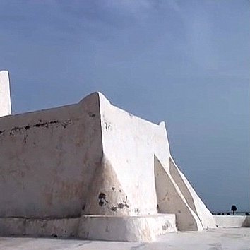 L'image montre un bâtiment de couleur blanche, typiquement de style architectural méditerranéen. Il semble être une petite structure, possédant une forme irrégulière avec des murs épais et angulaires. À son sommet, il y a une cloche ou un petit clocher. L'arrière-plan est plutôt clair, avec un ciel bleu ou nuageux qui met en valeur la luminosité de l'édifice. Ce type de construction pourrait évoquer des lieux historiques ou religieux, typiques de certaines régions côtières.