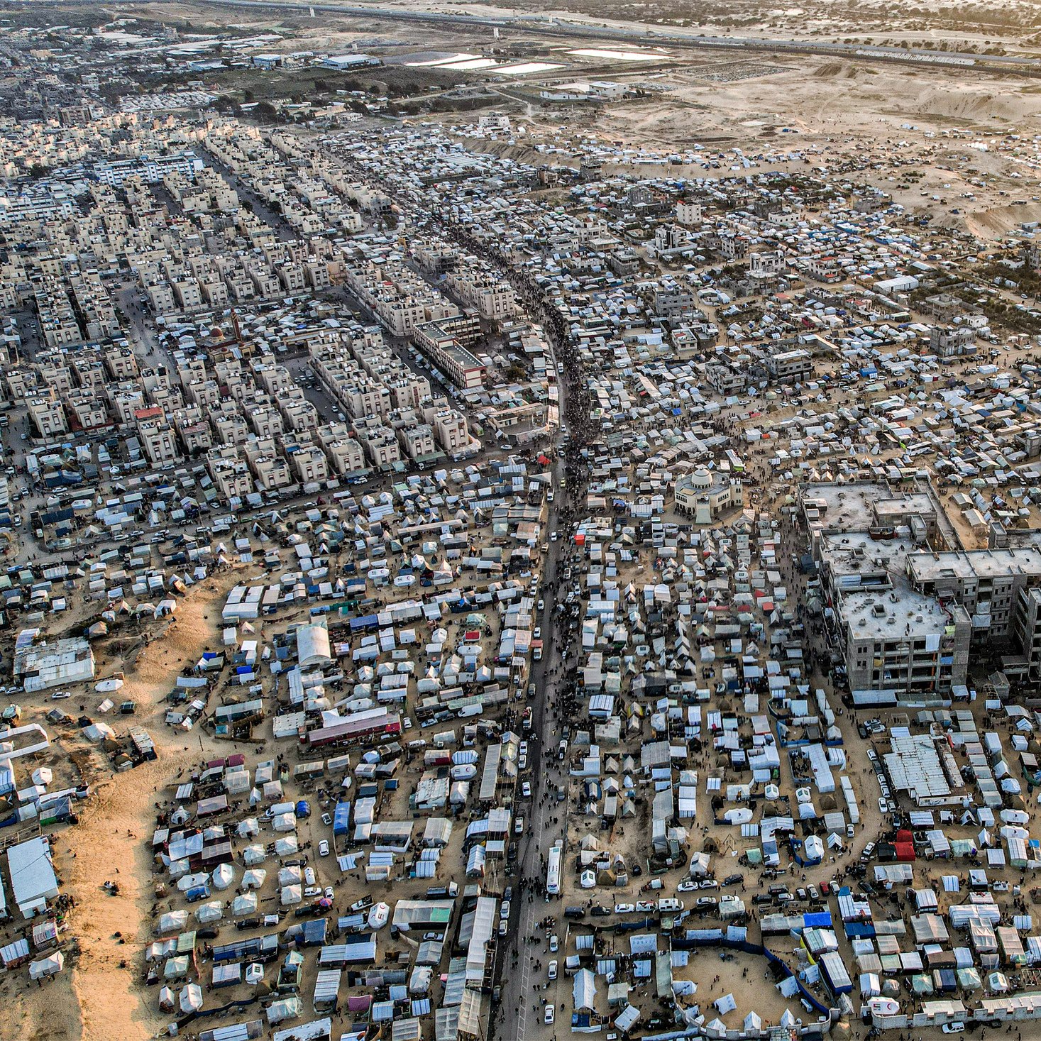 L'immagine mostra un'ampia area urbana vista dall'alto, caratterizzata da una fitta aggregazione di edifici e strutture. Si nota una forte densità abitativa, con molte abitazioni di dimensioni varie disposte in modo irregolare. Le strade principali e i passaggi sono visibili tra le varie costruzioni. In alcune zone si possono notare anche tende o strutture provvisorie, suggerendo la presenza di comunità temporanee. Sullo sfondo, si intravedono aree di terreni aperti e sviluppo urbano in espansione. La scena complessiva trasmette una sensazione di vivacità e organizzazione spontanea, con una tipologia abitativa che riflette una realtà complessa.