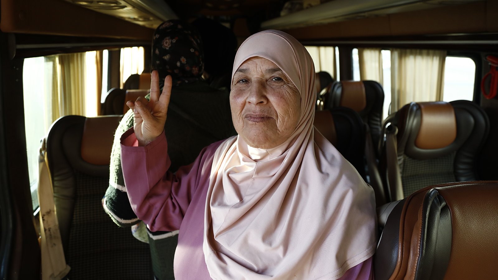 The image shows an elderly woman wearing a light pink hijab, smiling and making a peace sign with her right hand. She is inside a vehicle that appears to be a bus, with other passengers visible in the background. The interior of the bus features comfortable seating and soft lighting, contributing to a warm atmosphere. The woman's expression conveys friendliness and warmth.