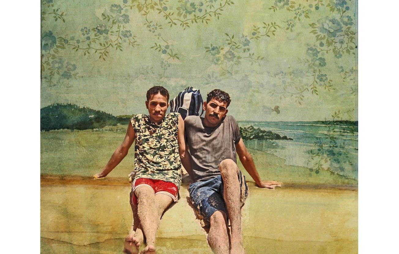 L'image représente deux jeunes hommes assis sur une plage. Ils portent des vêtements d'été ; l'un d'eux est en maillot de bain rouge et l'autre en shorts. Leur posture décontractée et le fond de plage évoquent une ambiance estivale. En arrière-plan, on peut voir des éléments naturels tels que l'eau et un paysage côtier, le tout stylisé par une texture artistique et des motifs floraux.