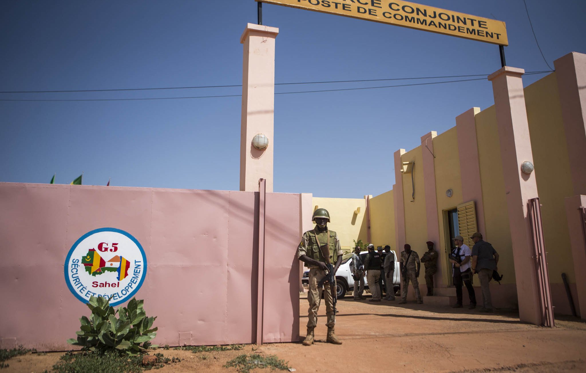 L'image montre un bâtiment qui semble être un poste de commandement de la "Force Conjointe" au sein du G5 Sahel. À l'entrée, on voit un mur avec le logo du G5, ainsi qu'un panneau indiquant "FORCE CONJOINTE POSTE DE COMMANDEMENT". Un soldat en uniforme est en position de garde près de l'entrée, tandis que d'autres personnes sont visibles à l'intérieur ou autour du bâtiment. L'environnement est désertique, et le ciel est dégagé.