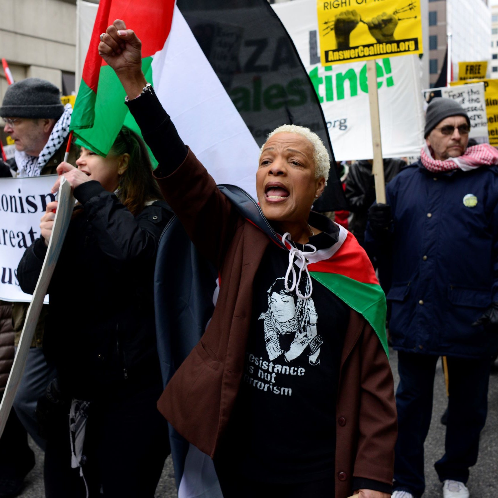 L'image montre une manifestation avec un groupe de personnes brandissant des pancartes et des drapeaux. Une femme au premier plan, portant un t-shirt avec une inscription, lève le poing en criant des slogans. Les manifestants semblent exprimer leur soutien à la cause palestinienne. On peut apercevoir des drapeaux aux couleurs palestiniennes, ainsi que des affiches dénonçant le sionisme et appelant à la résistance. L'ambiance semble dynamique et engagée.