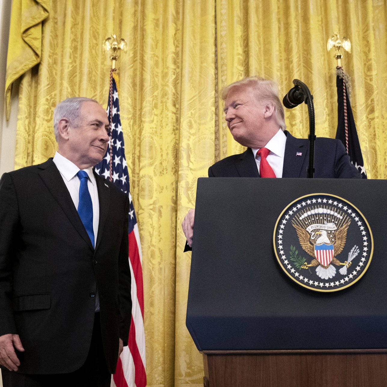 L'image montre deux hommes debout devant un podium avec le sceau présidentiel. En arrière-plan, il y a des rideaux en velours doré et des drapeaux, dont un drapeau américain. Les deux hommes échangent un regard complice, suggérant une atmosphère amicale. L'un est en costume sombre et l'autre porte une cravate rouge, ce qui attire l'attention. L'environnement semble formel, probablement lors d'un événement officiel.