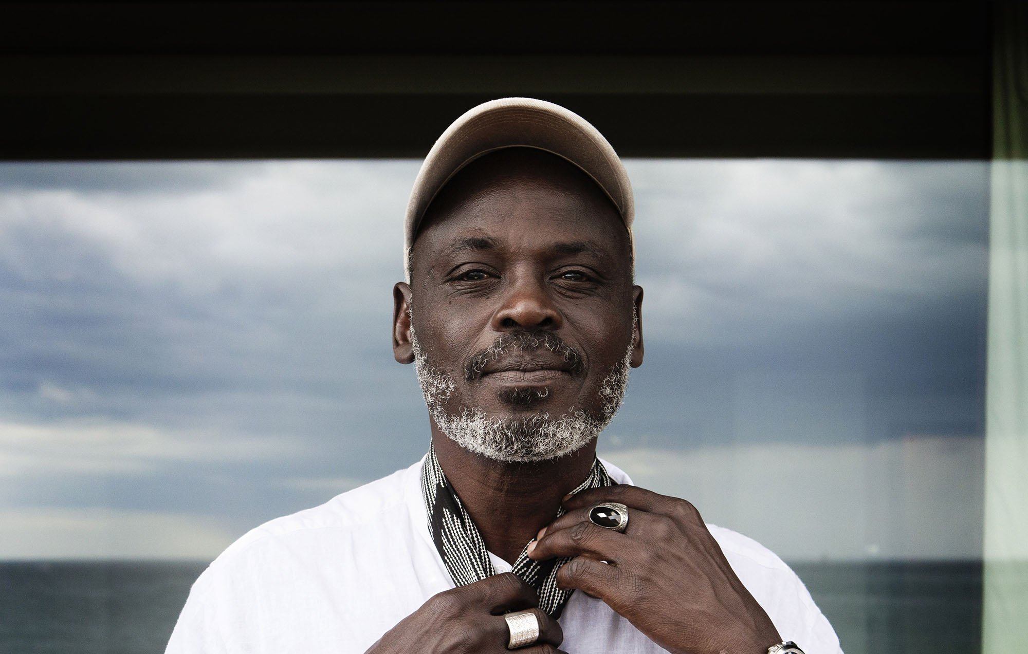 L'image montre un homme debout, portant une chemise blanche et un chapeau beige. Il est en train de nouer une écharpe autour de son cou. Son visage est marqué par l'âge, avec une barbe grise bien entretenue. Il porte également plusieurs bagues aux doigts et une montre en cuir. En arrière-plan, on peut voir une fenêtre qui donne sur une mer calme, sous un ciel nuageux. L'ensemble de l'image dégage une atmosphère tranquille et contemplative.