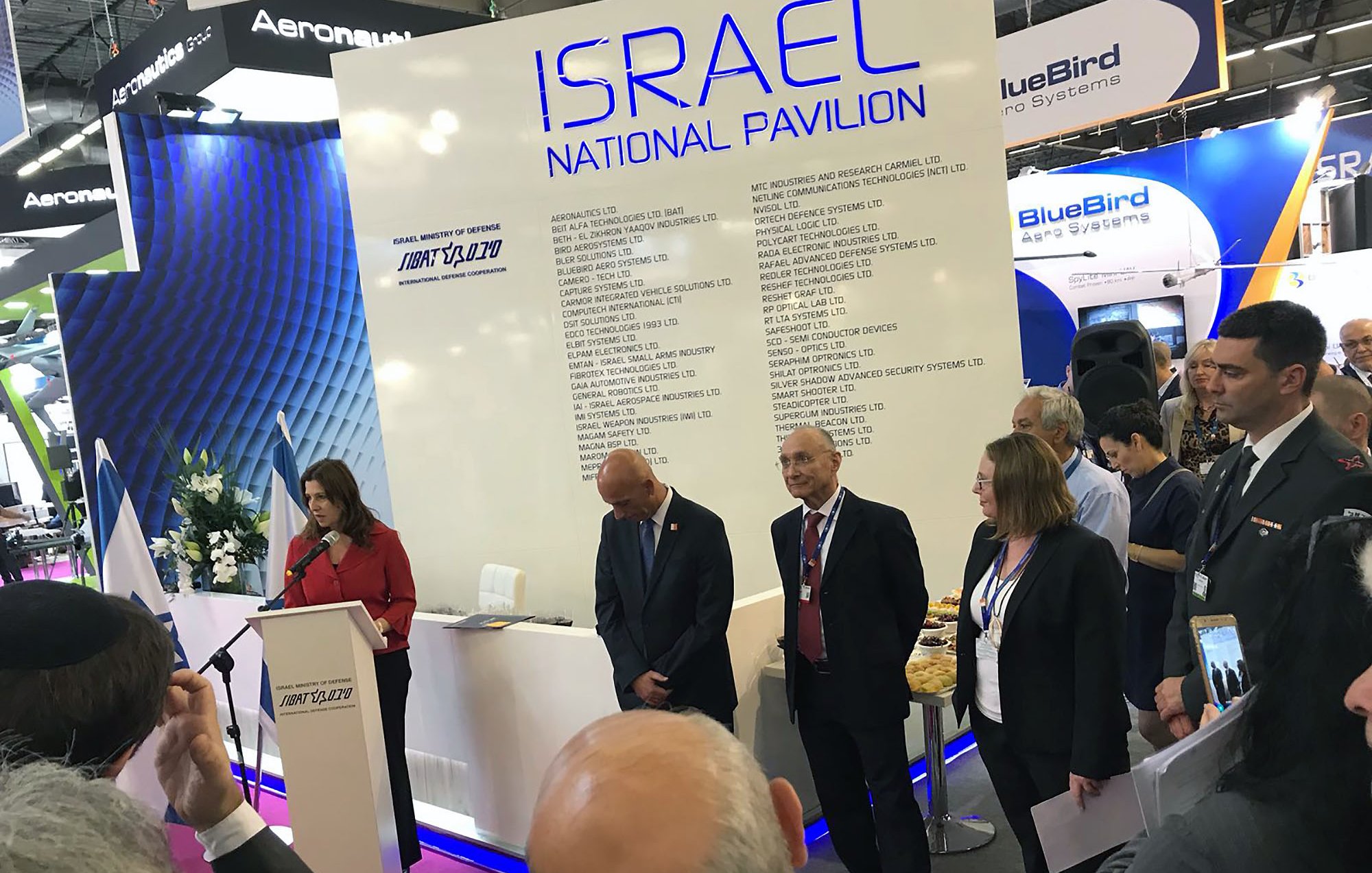 تظهر الصورة مجموعة من الأشخاص في حدث أو معرِض، حيث يتواجد متحدثة أمام منصة تحمل شعار "إسرائيل" وتفاصيل عن الجناح الوطني. هناك بعض الحضور في الخلفية يستمعون بانتباه، بينما يبدو بعضهم منشغلاً بتدوين الملاحظات. المنصة محاطة بالزهور، ويوجد مرافقون يرتدون بدلات رسمية. الأجواء تعكس نشاطًا رسميًا ومهنيًا.