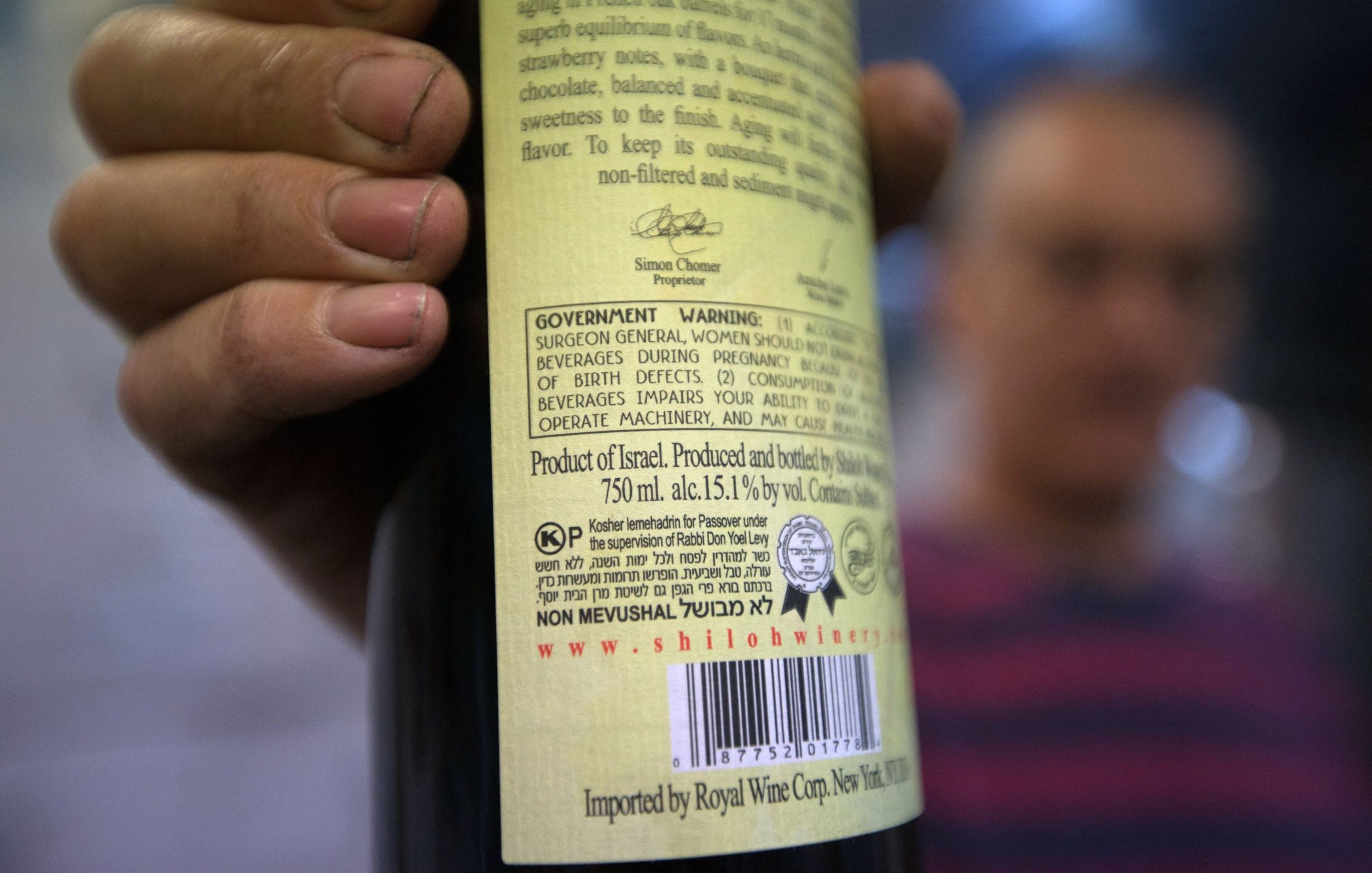 L'image montre une personne tenant une bouteille de vin, avec l'étiquette clairement visible. L'étiquette comporte des informations détaillées sur le produit, y compris des avertissements et des mentions sur la provenance, indiquant qu'il s'agit d'un vin produit en Israël. La bouteille est de 750 ml et a un taux d'alcool de 15,1 % vol. En arrière-plan, on peut deviner une autre personne floue, mais les détails ne sont pas très clairs. Les mains de la personne tenant la bouteille semblent légèrement sales, ce qui pourrait suggérer un environnement de travail ou une activité en plein air.