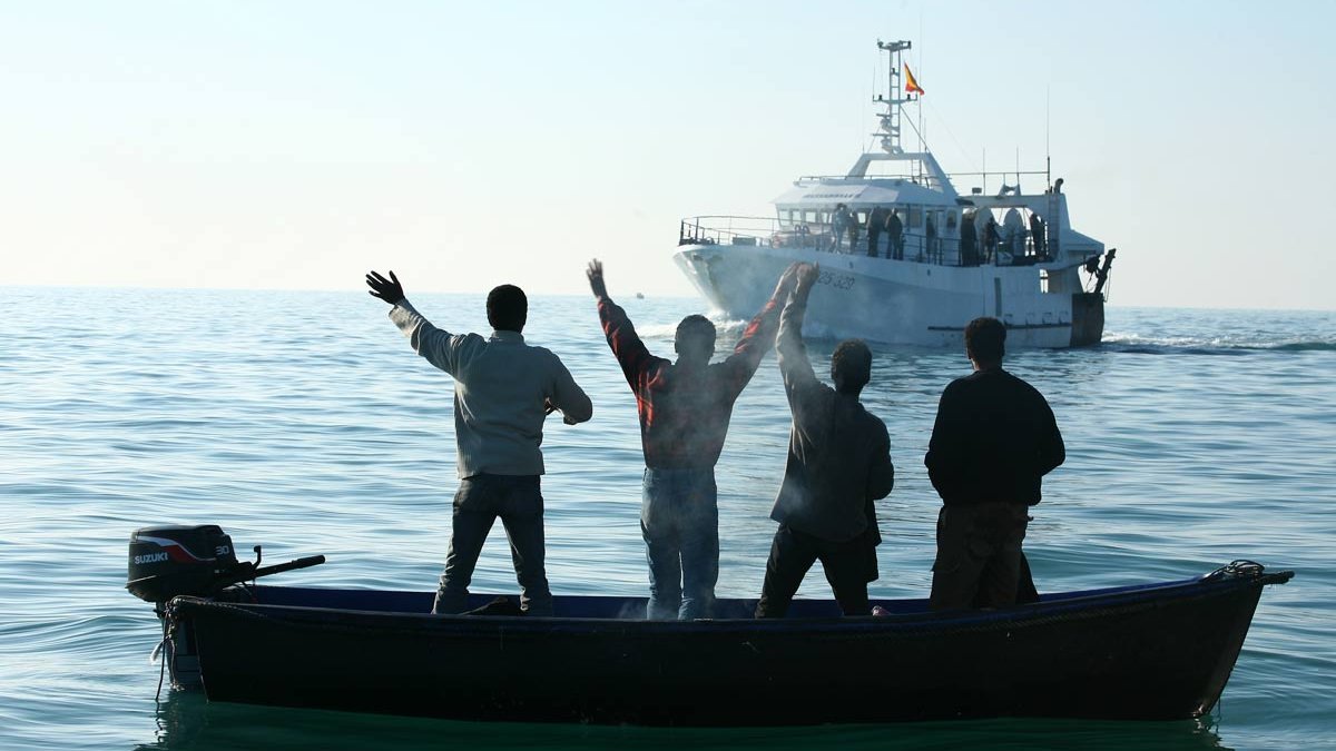 L'image montre un groupe de quatre personnes se tenant sur un petit bateau. Elles sont de dos et lèvent les bras en signe d'accueil ou de victoire. En arrière-plan, un grand bateau navigue à proximité dans des eaux calmes, baignant dans une lumière douce. L'ambiance semble joyeuse et pleine d'enthousiasme.
